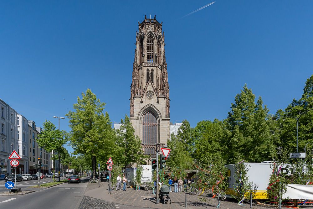 Große Kirche im gotischen Stil an einer Straße mit mehreren Bäumen und Fußgängern im Vordergrund.