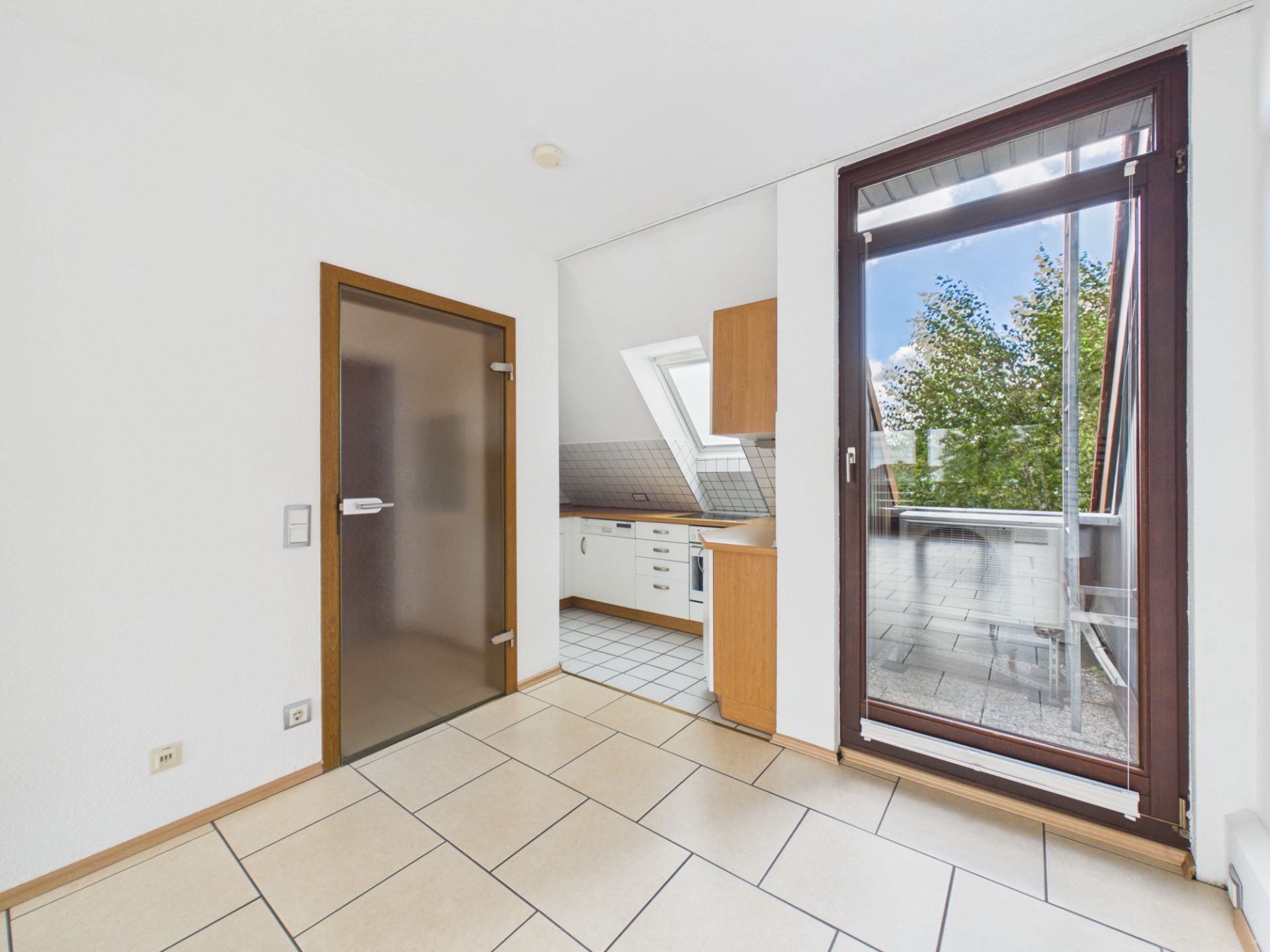 Heller Raum mit geflißtem Boden, Zugang zur Küche und Glastür zur Terrasse.