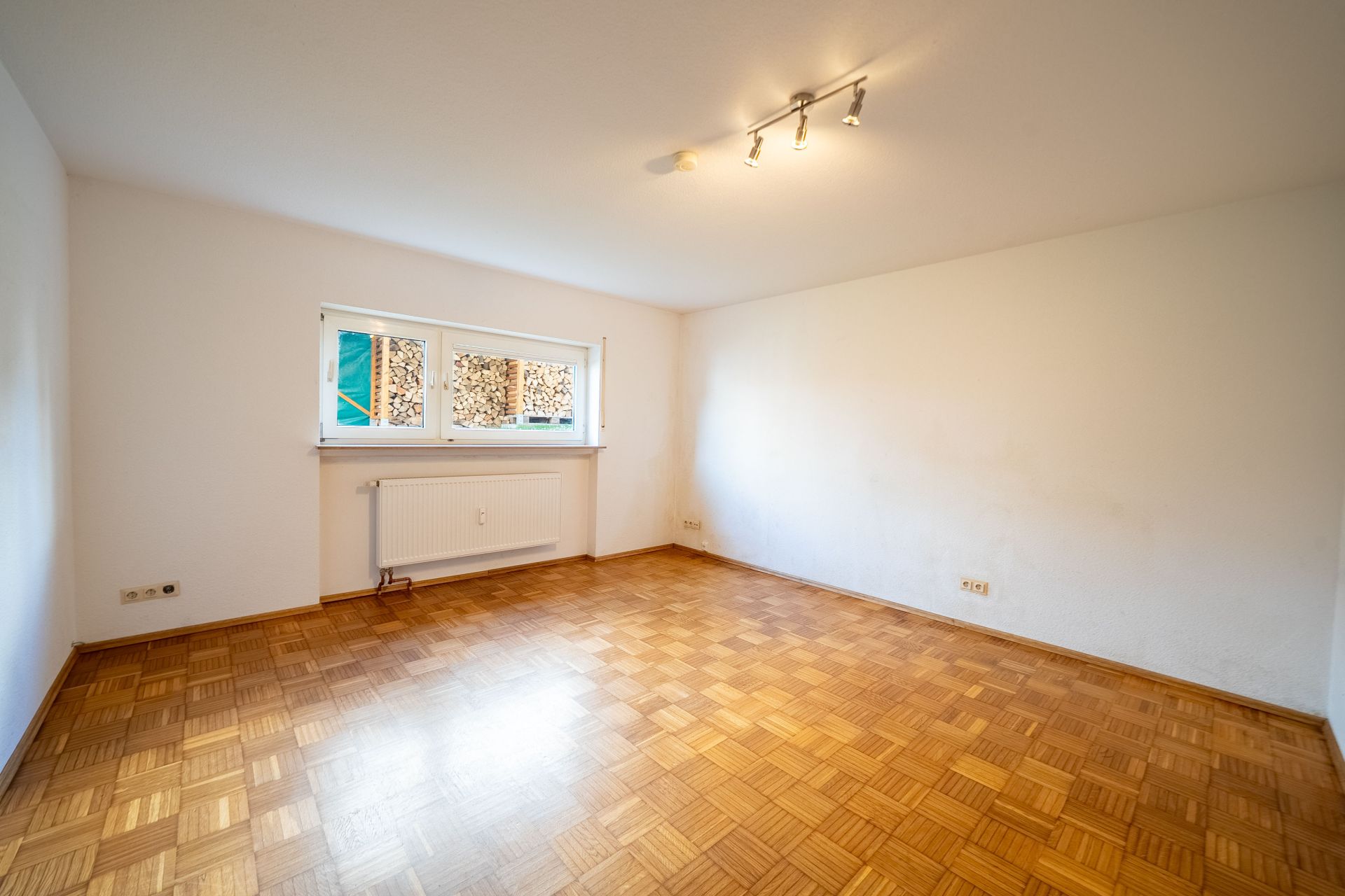 Leeres Zimmer mit Parkettboden, weißen Wänden, Fenster und Deckenleuchte.
