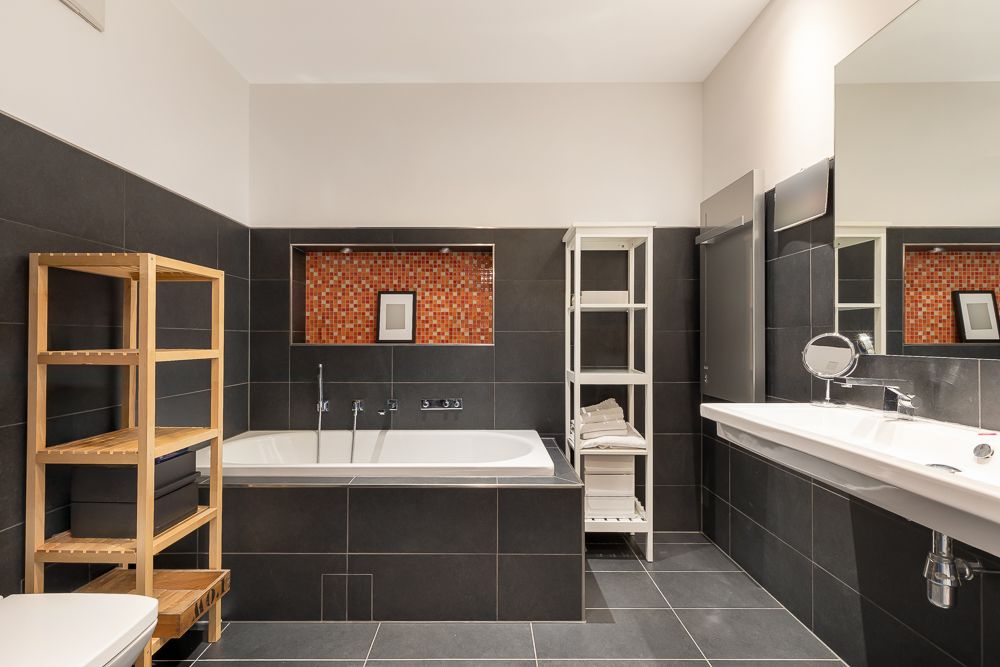 Modernes Badezimmer mit Badewanne, Regal, Waschbecken und dunklen Fliesen.