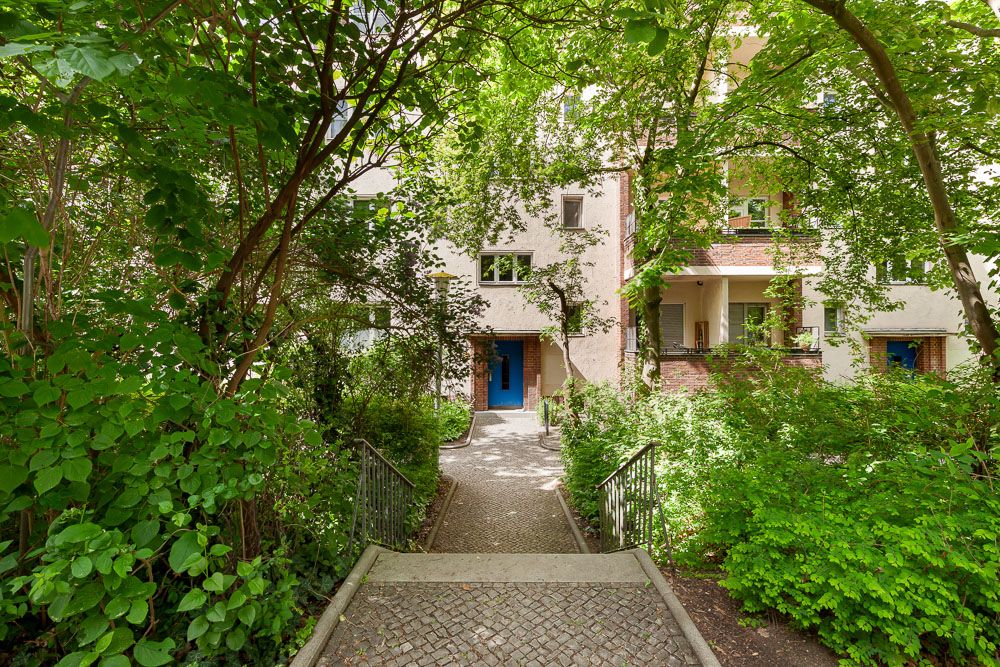 Weg mit Geländer führt durch grünen Garten zu einem Mehrfamilienhaus mit blauen Türen.