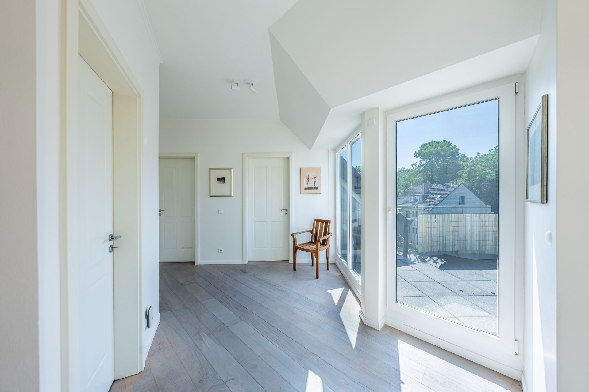Heller Flur mit Holzboden, weißen Türen, großem Fenster und einem Stuhl neben der Terrassentür.