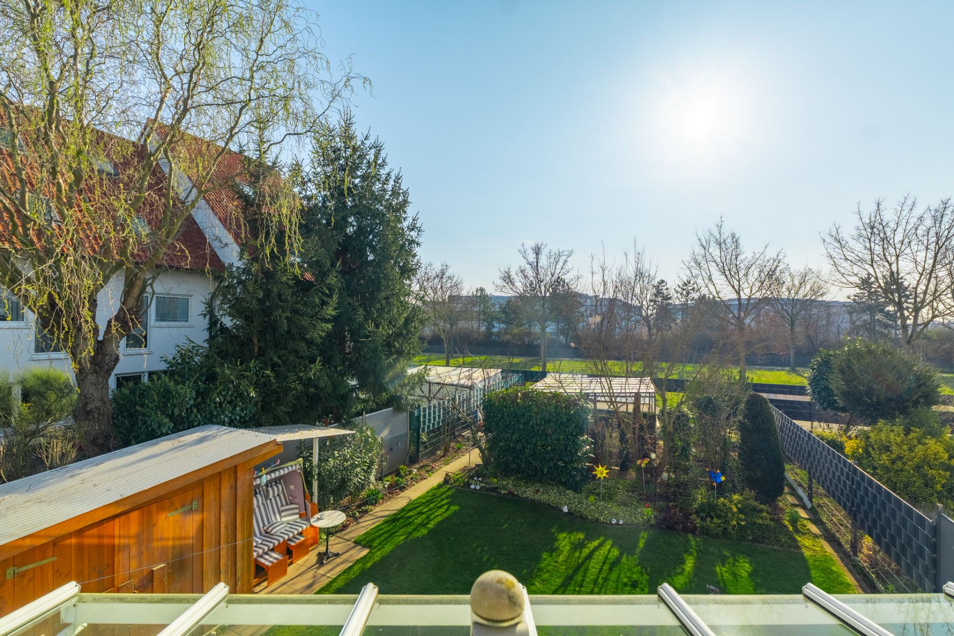Blick auf einen sonnigen Garten mit Rasen, Bäumen und einer Holzbank von einem Balkon aus.