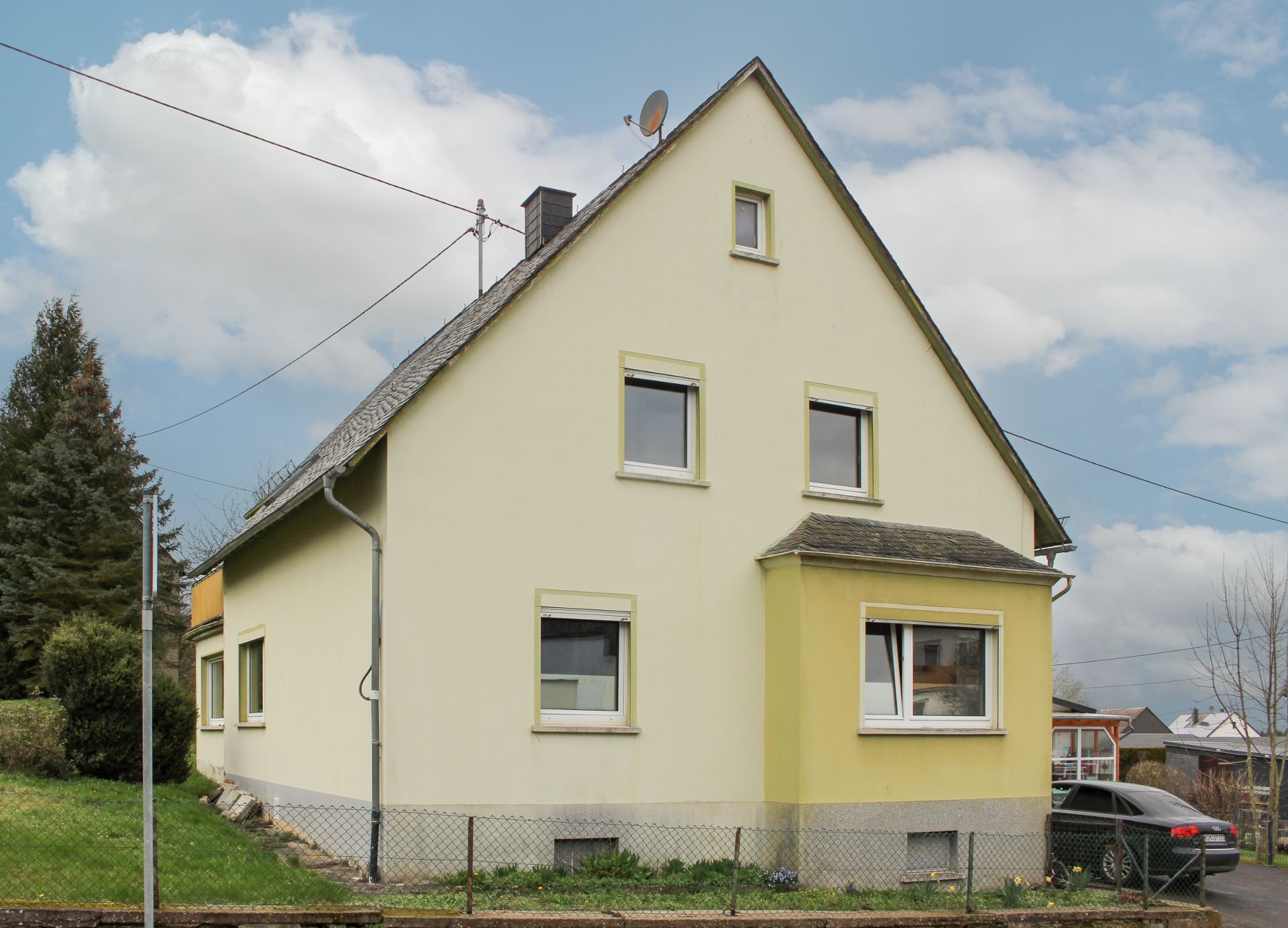 Freistehendes, gelbes Einfamilienhaus mit Satteldach und Gartenzaun, Auto links im Bild.