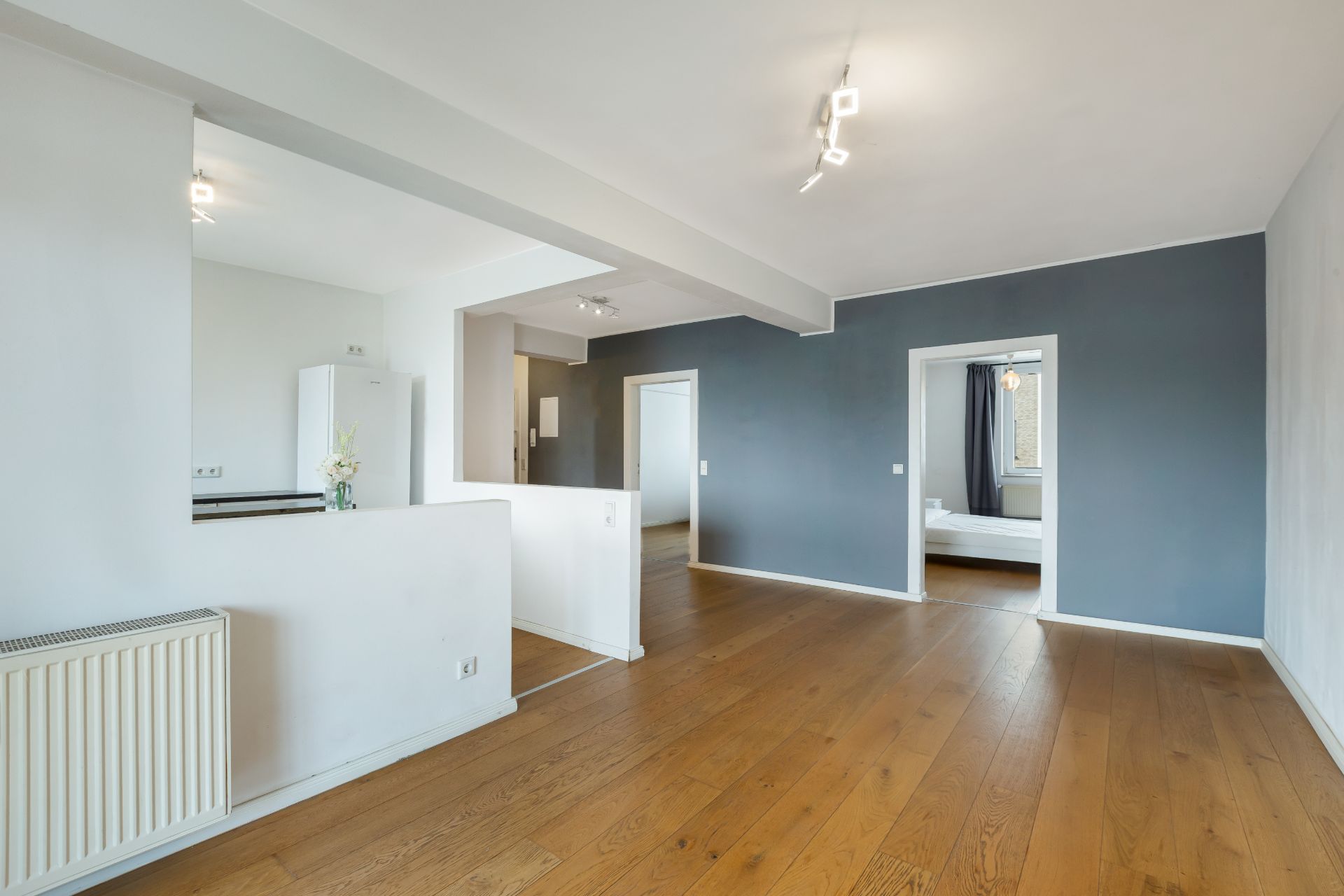Helles Wohnzimmer mit Holzboden, grauer Wand und Zugang zu einem Schlafzimmer.