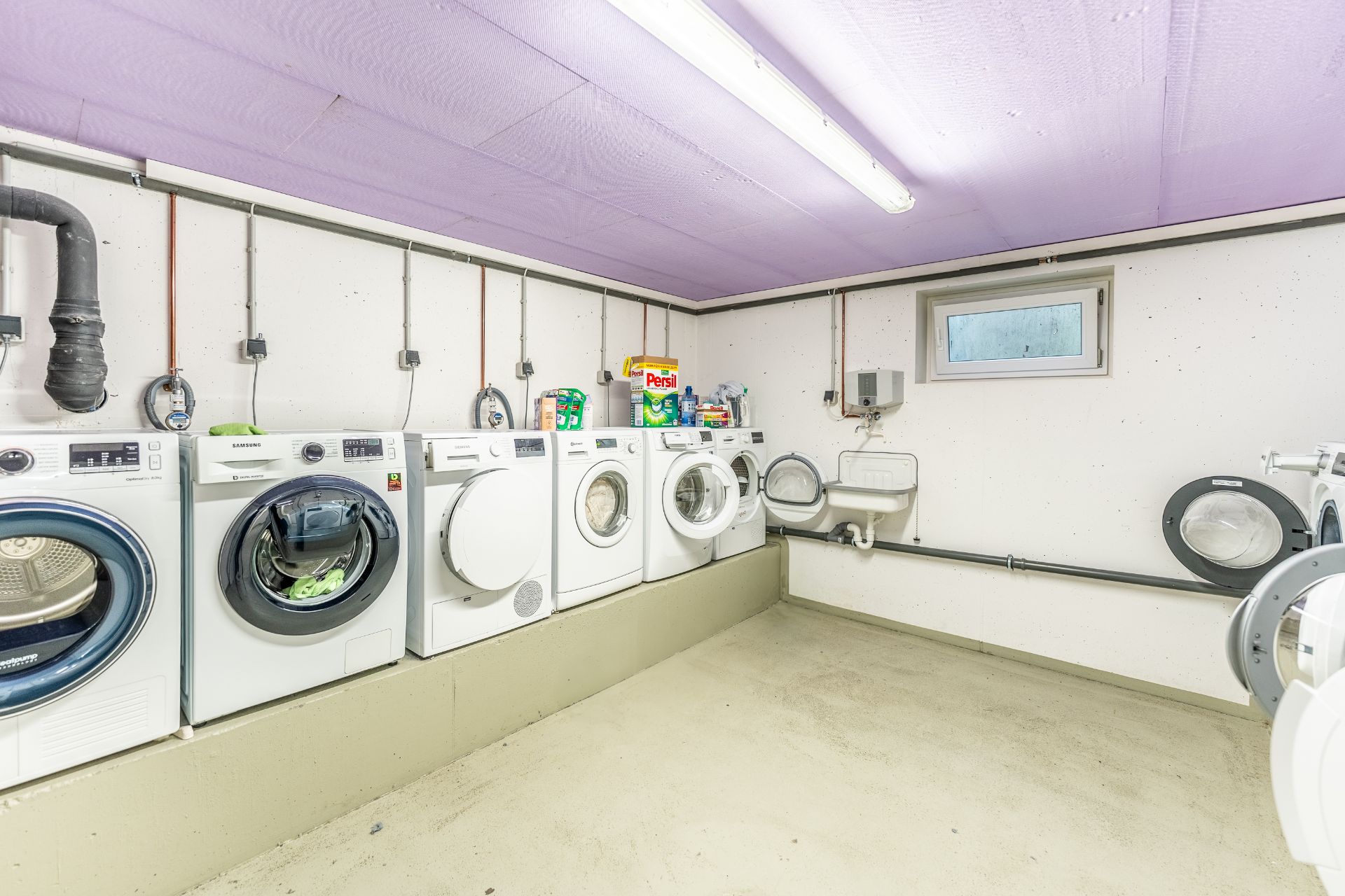 Waschraum mit mehreren Waschmaschinen, Trocknern, Waschbecken und einem kleinen Fenster.