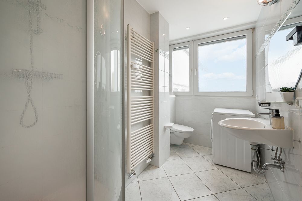 Helles Badezimmer mit Dusche, Waschbecken, Toilette, Waschmaschine und großem Fenster.