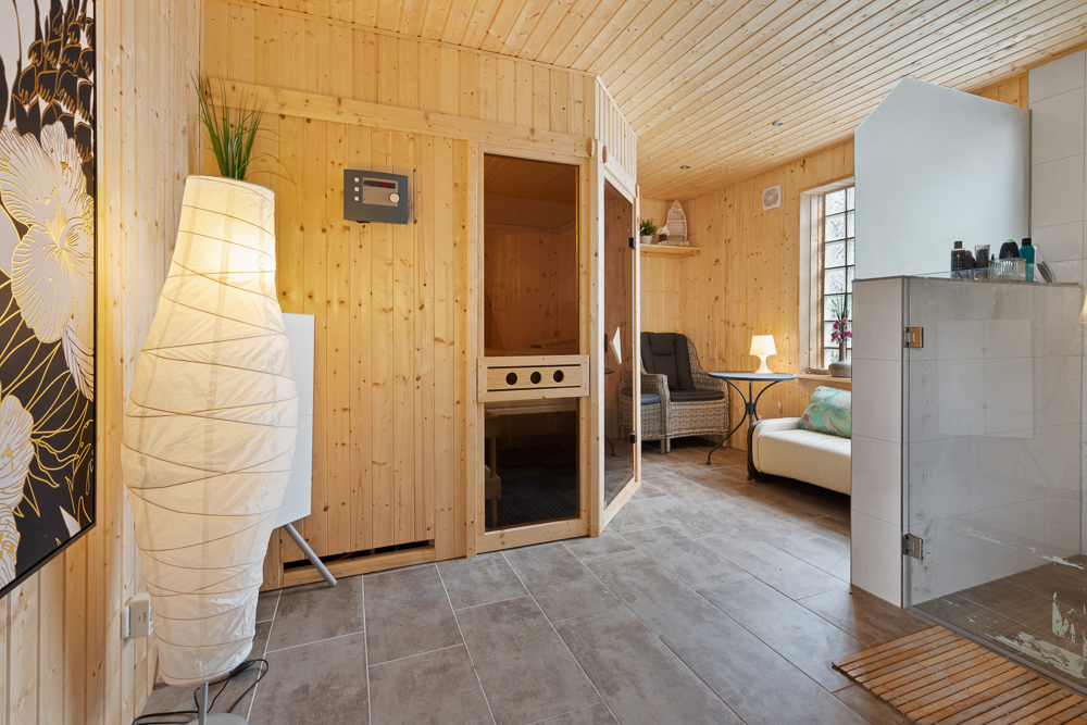 Heller Wellnessraum mit Sauna, Duschkabine, Stehlampe und Sitzbereich aus Holz.