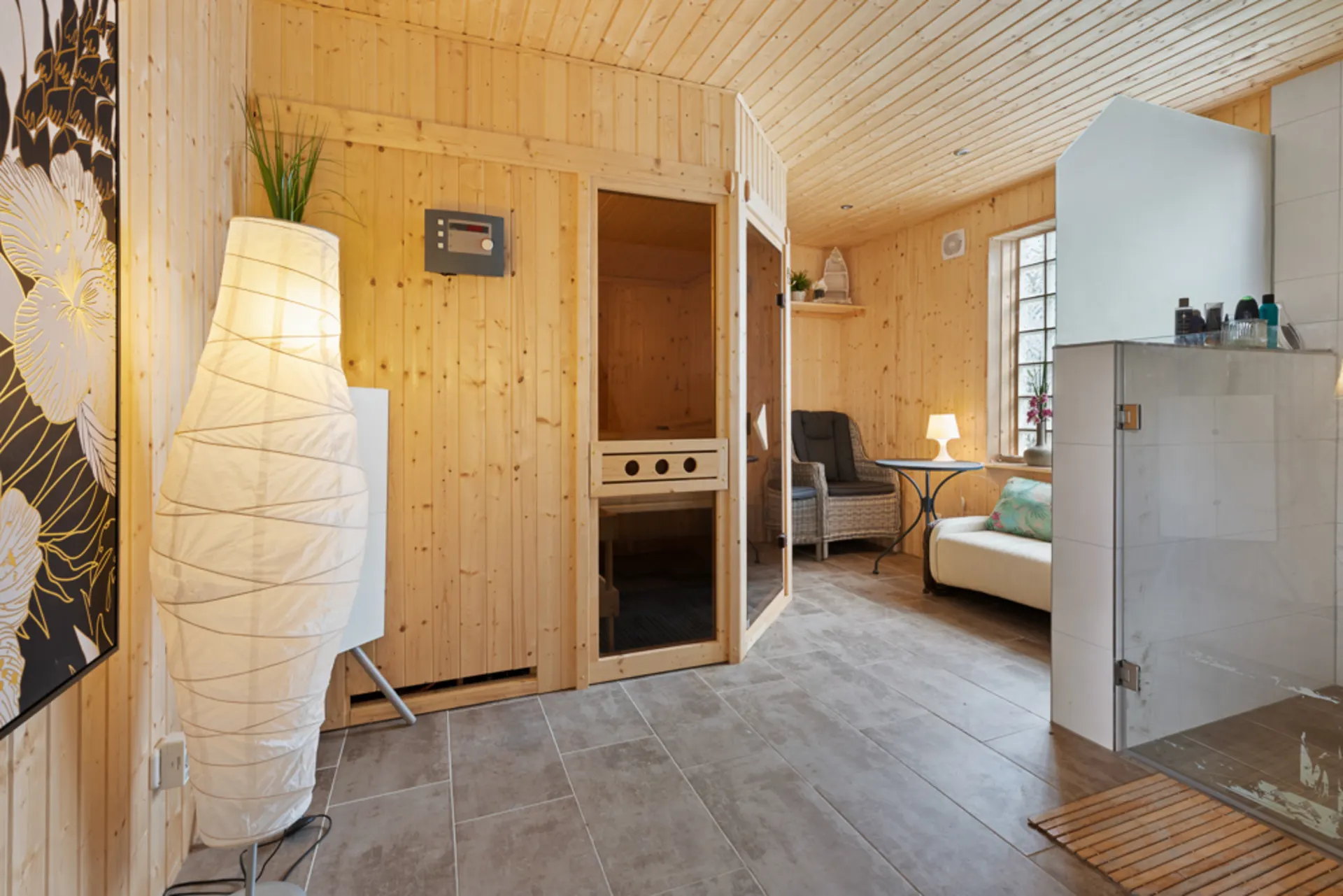 Heller Wellnessraum mit Sauna, Duschkabine, Stehlampe und Sitzbereich aus Holz.