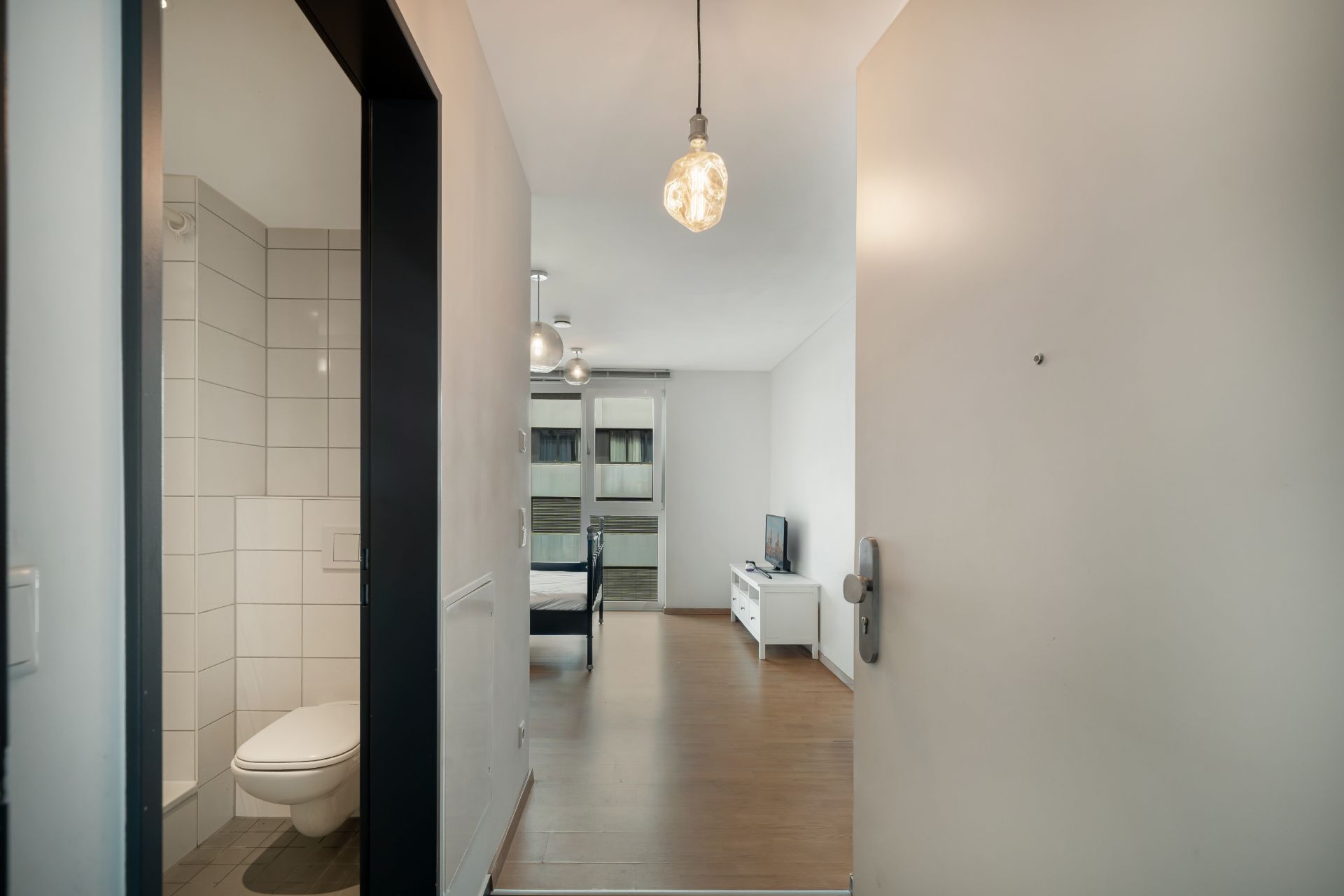 Blick in einen Flur mit Zugang zu einem Bad und einem Wohnraum mit Bett und Fernseher.