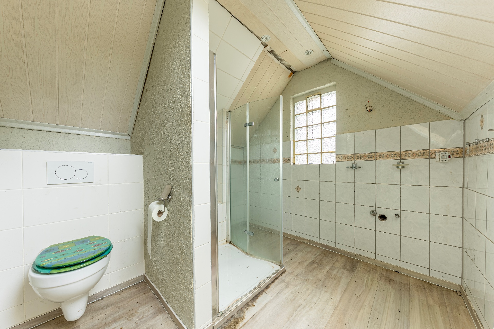 Helles Badezimmer mit schräger Decke, Dusche, Wandtoilette und gefliestem Bereich.