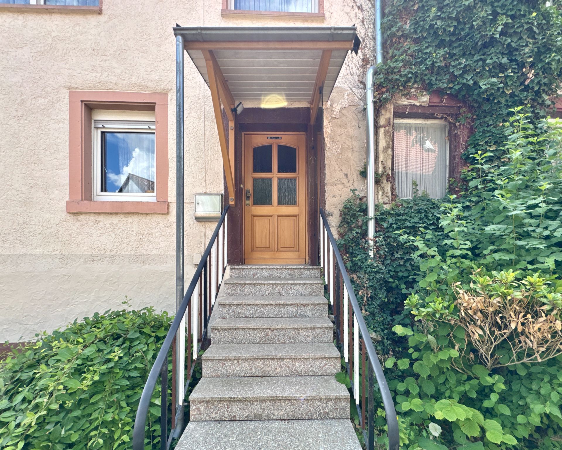 Eingangstür mit Überdachung, Treppe und viel Grün neben dem Haus.