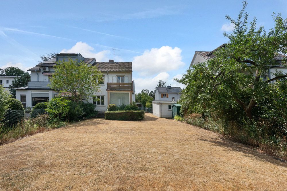 Ein Wohnhaus mit großem, trockenem Garten und umliegenden Bäumen bei sonnigem Wetter.