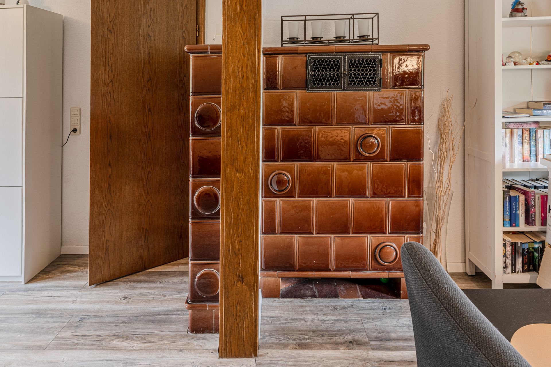 Brauner Kachelofen neben Holztür, mit Regal und Stuhl im Wohnzimmer.