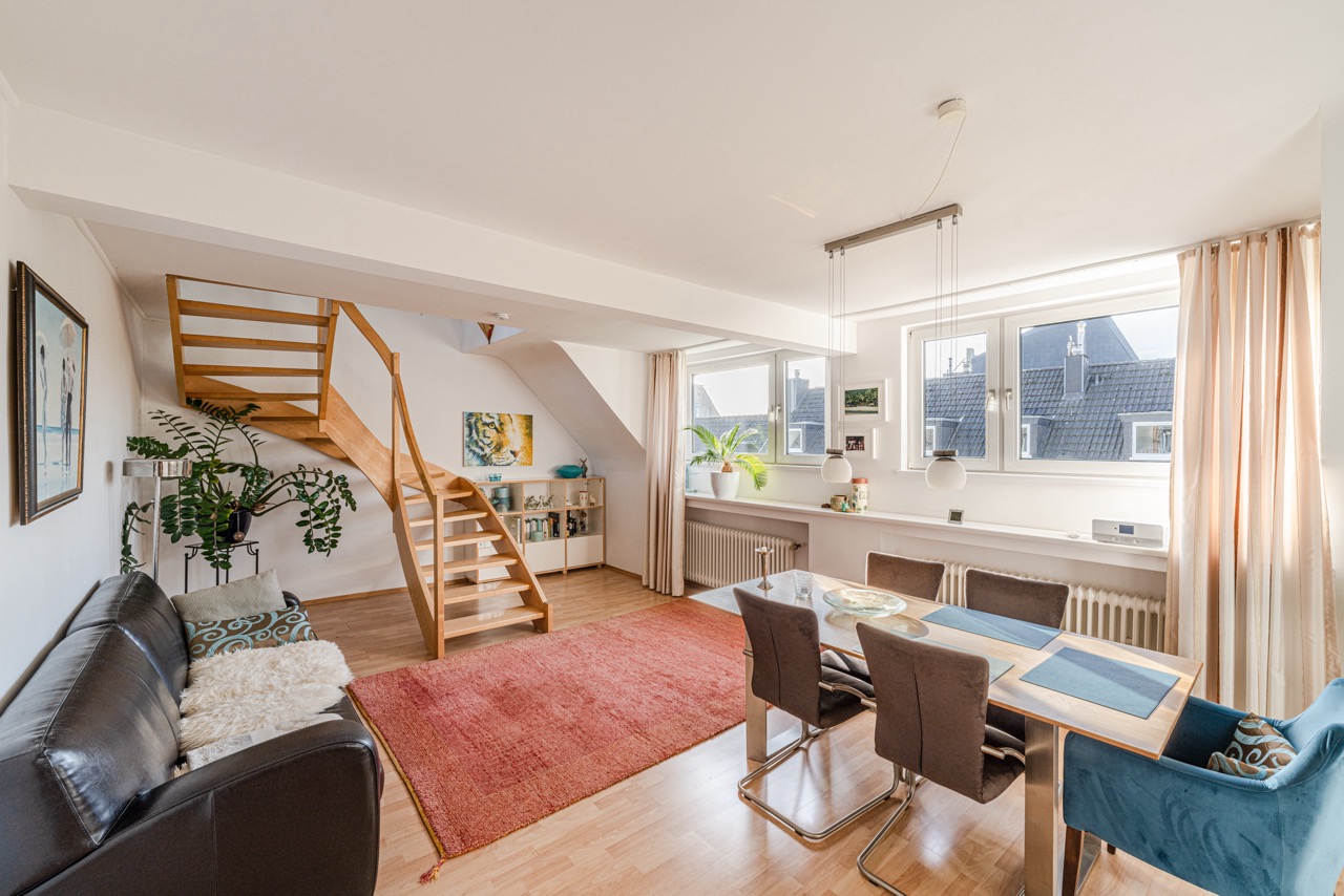 Helles Wohnzimmer mit Holztreppe, Sofa, Esstisch, Teppich und großen Fenstern.