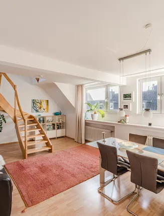 Helles Wohnzimmer mit Holztreppe, Sofa, Esstisch, Teppich und großen Fenstern.