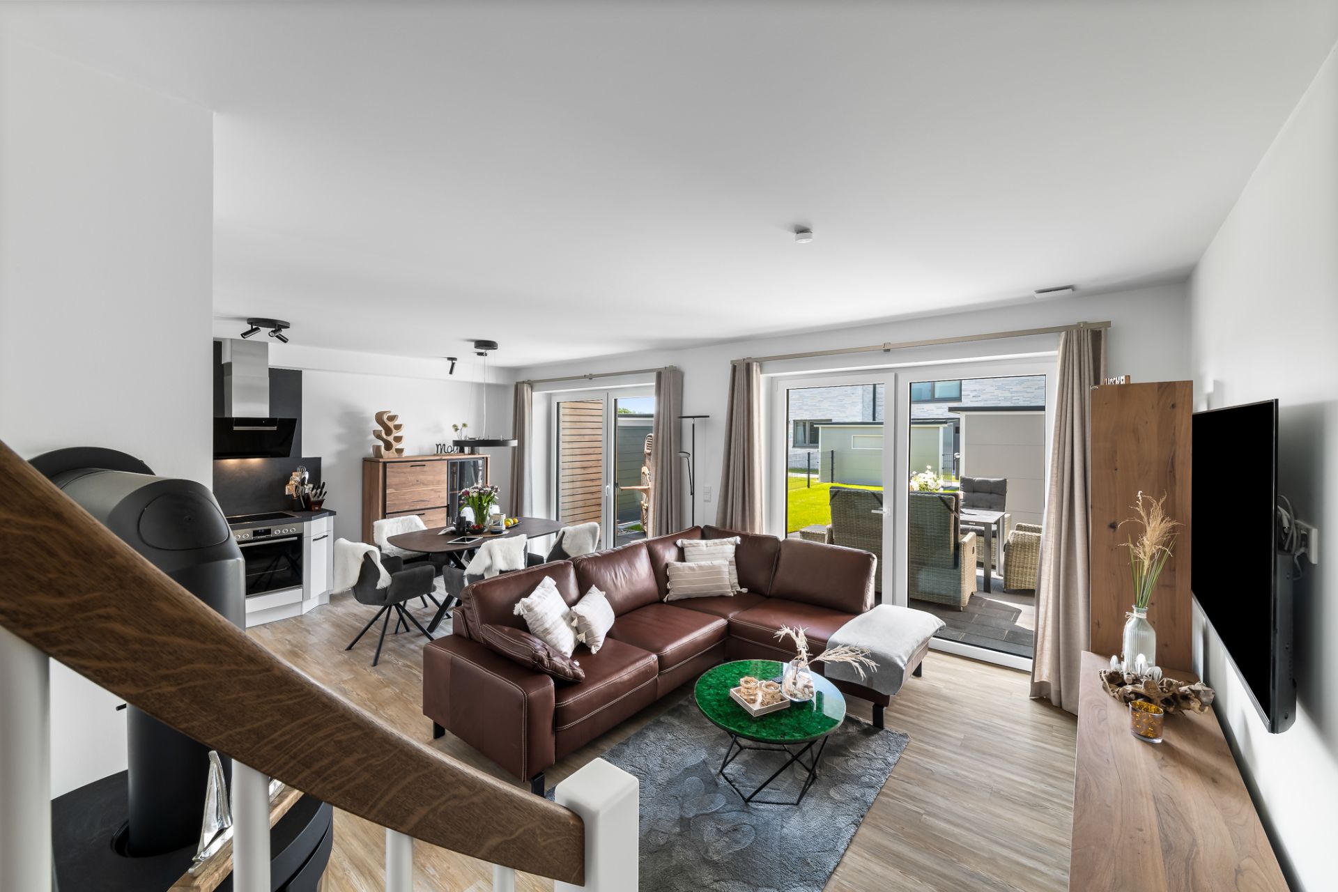 Modernes Wohnzimmer mit braunem Sofa, Esstisch, Küche und Zugang zur Terrasse.