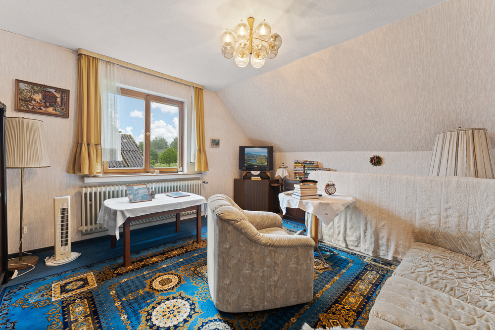 Wohnzimmer mit blauem Teppich, Sofa, Sessel, Tisch, Fernseher und Fenster mit gelben Vorhängen.