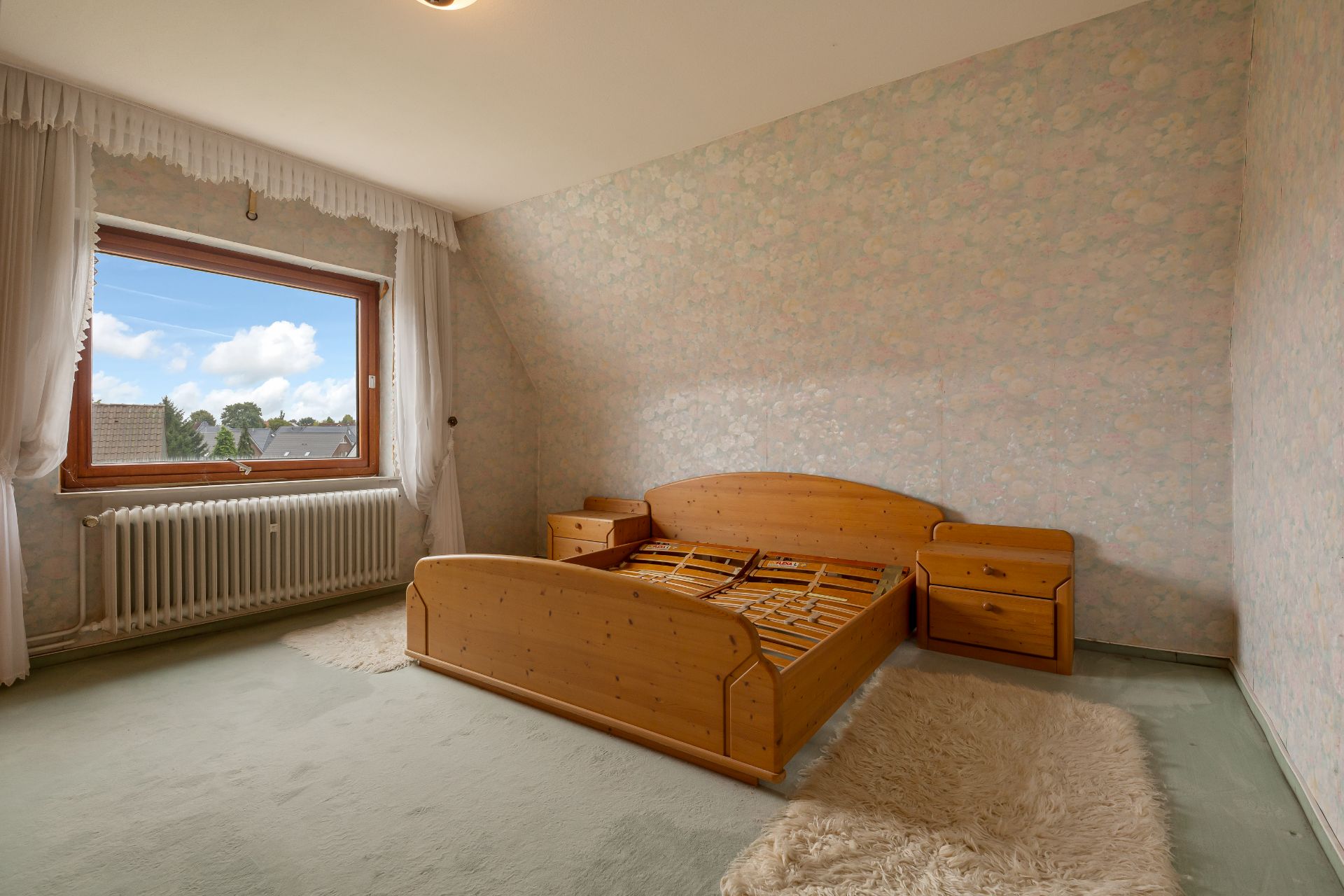 Leeres Schlafzimmer mit Holzrahmenbett, zwei Nachttischen, Teppich und großem Fenster.