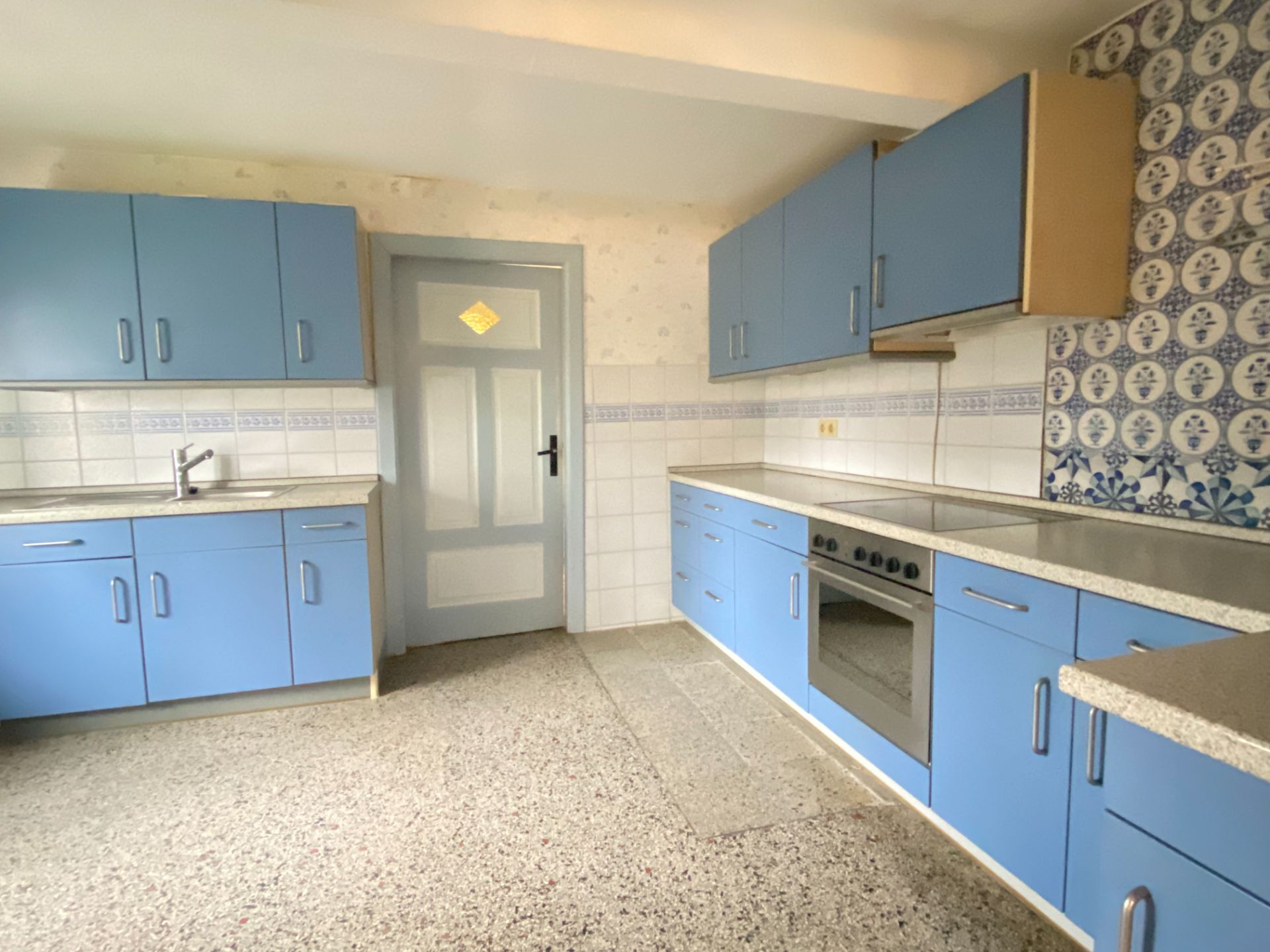 Helle Küche mit blauen Schränken, Elektroherd und gemusterten Fliesen an einer Wand.