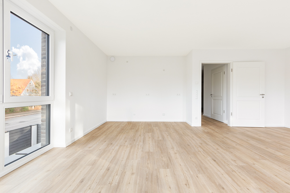 Helles, leeres Zimmer mit großen Fenstern, Holzboden und weißen Wänden.