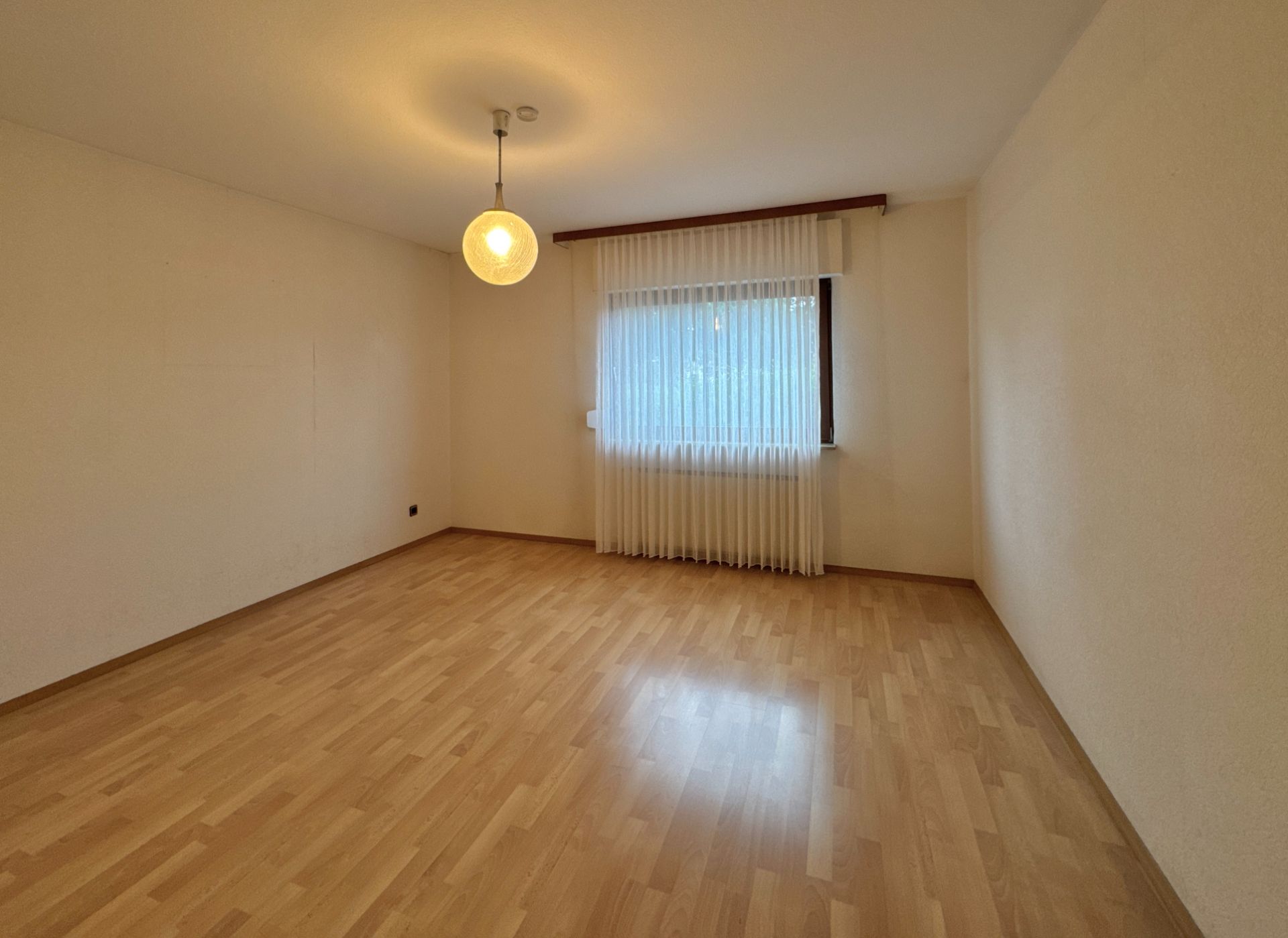 Leerer Raum mit hellem Laminatboden, Fenster mit Gardine und Deckenleuchte.