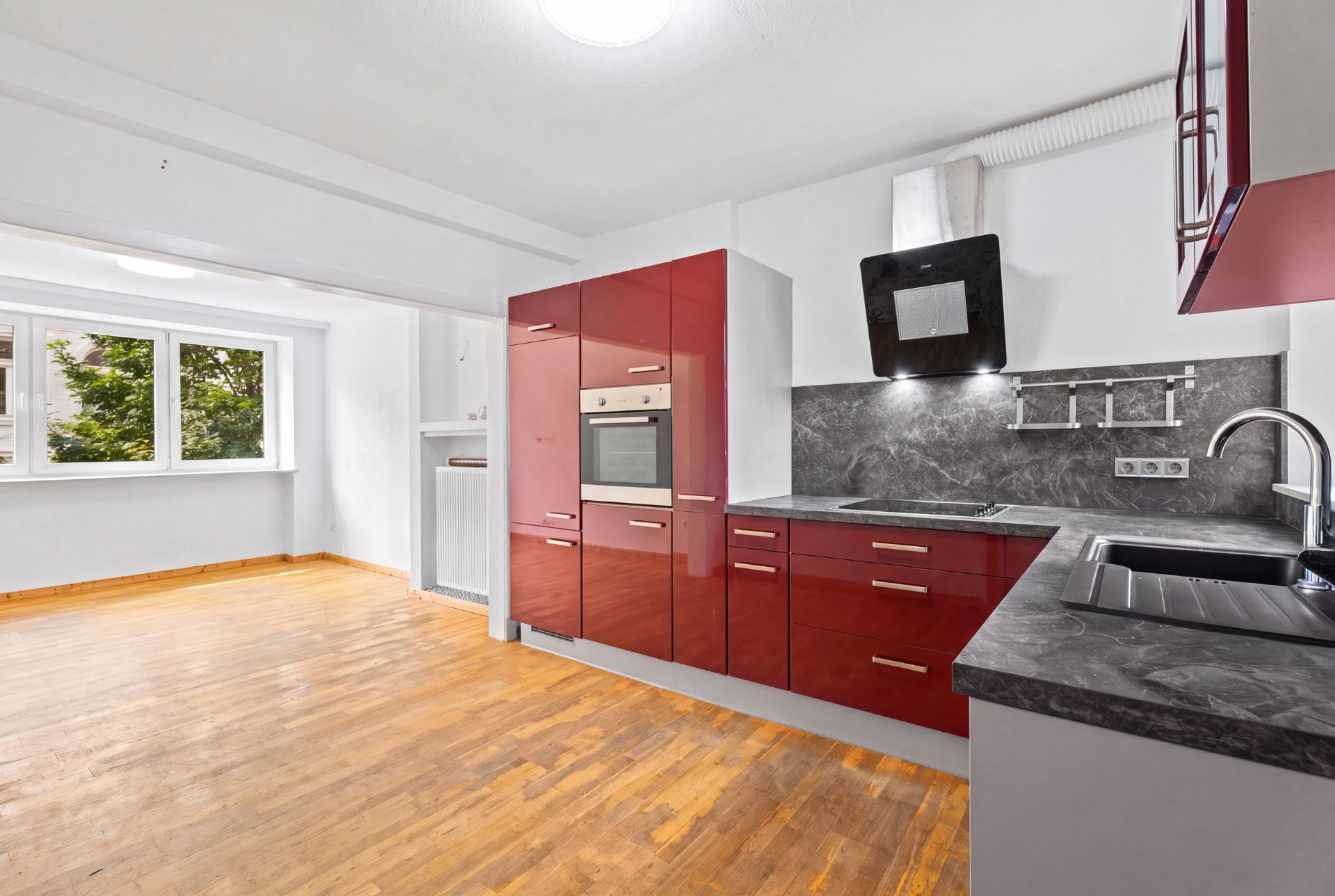 Moderne Küche mit roten Hochglanzfronten und Holzfußboden, daneben ein heller Essbereich mit Fenstern.
