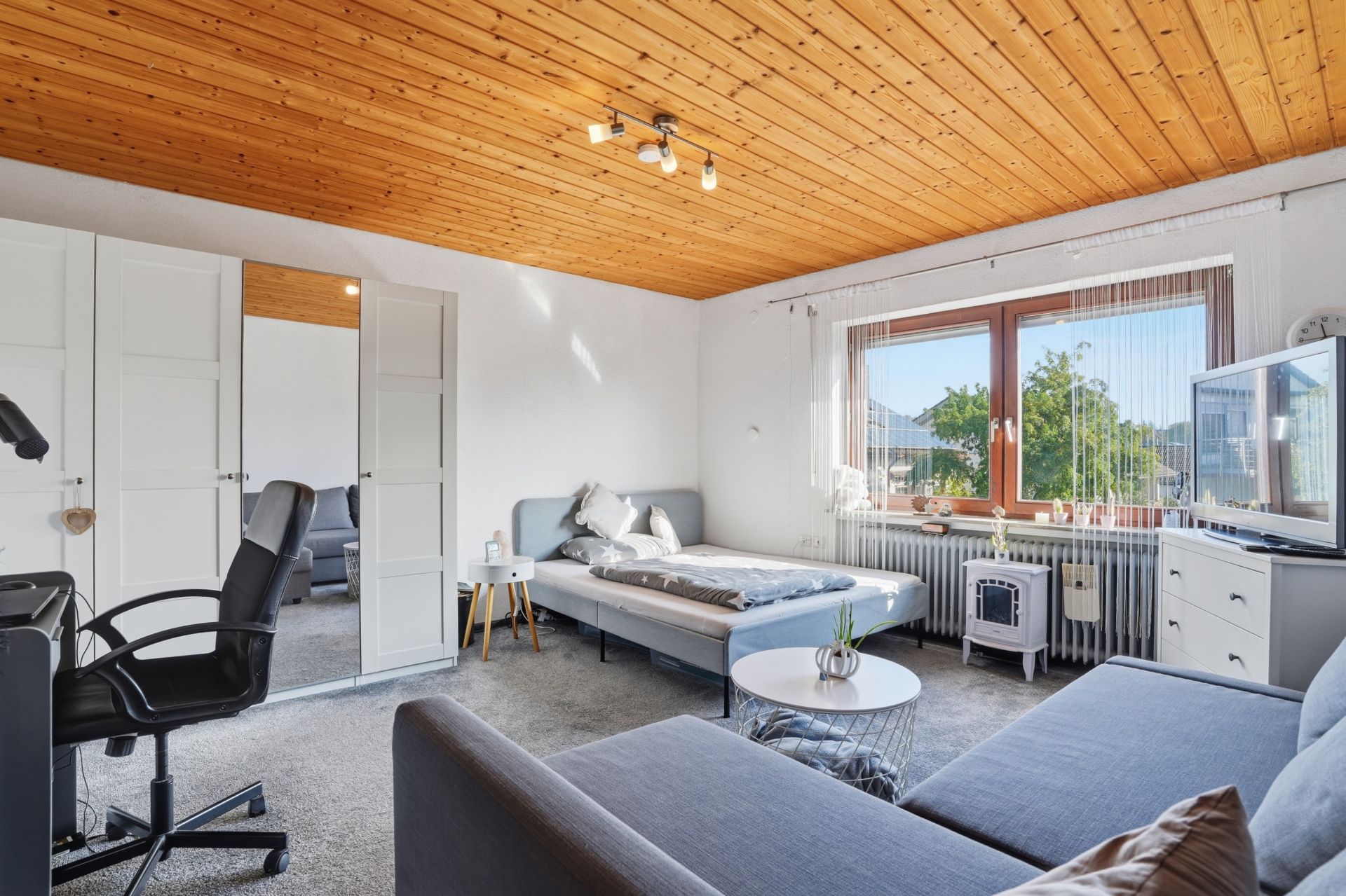 Helles Wohnzimmer mit Holzbalkendecke, Bett, grauem Sofa, Schreibtisch und großem Fenster.