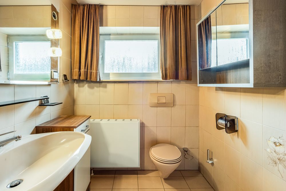 Helles Badezimmer mit Waschbecken, Toilette, Spiegelschrank und zwei Fenstern mit Vorhängen.