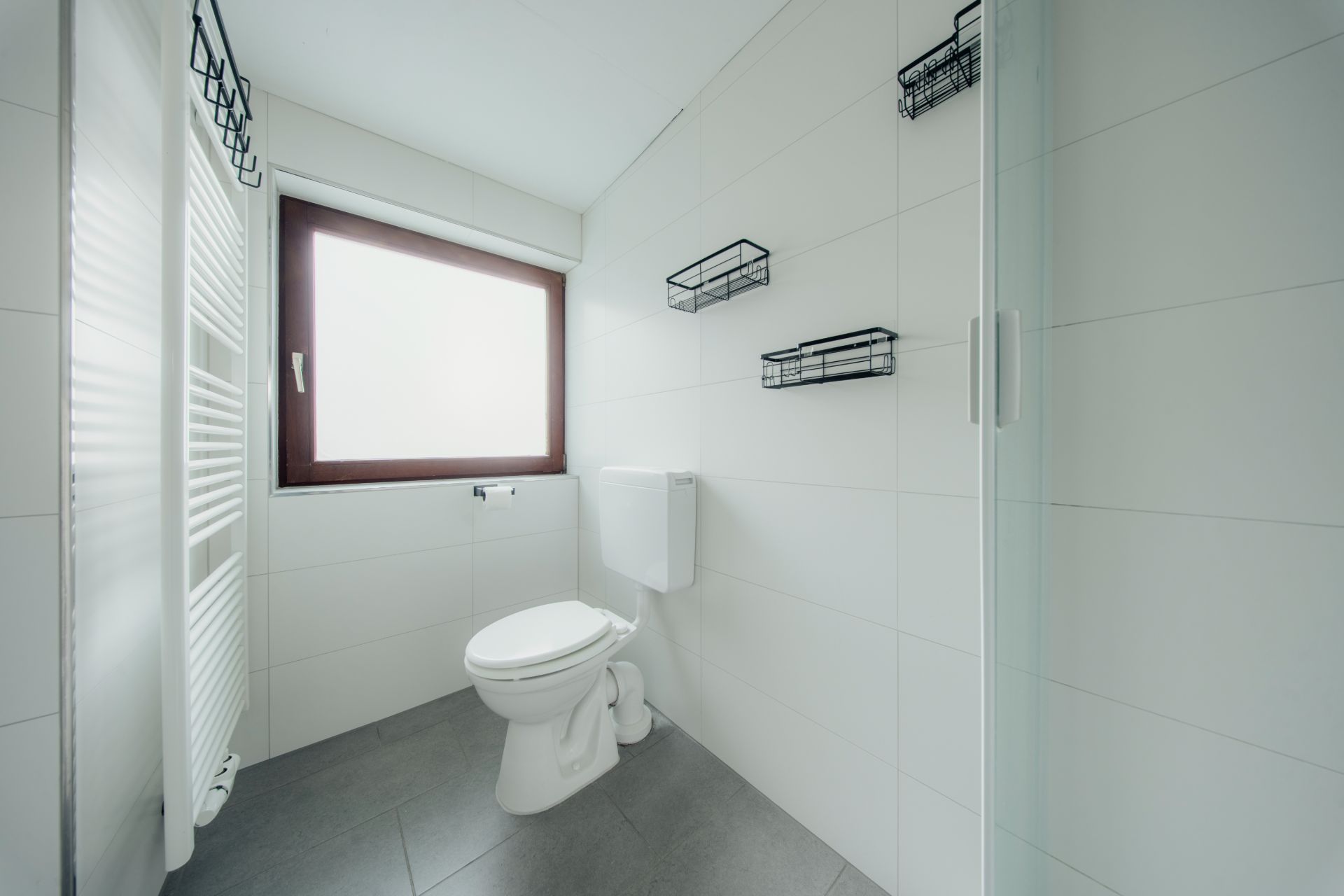 Helles Badezimmer mit Toilette, Fenster, Handtuchheizkörper und drei Wandregalen.