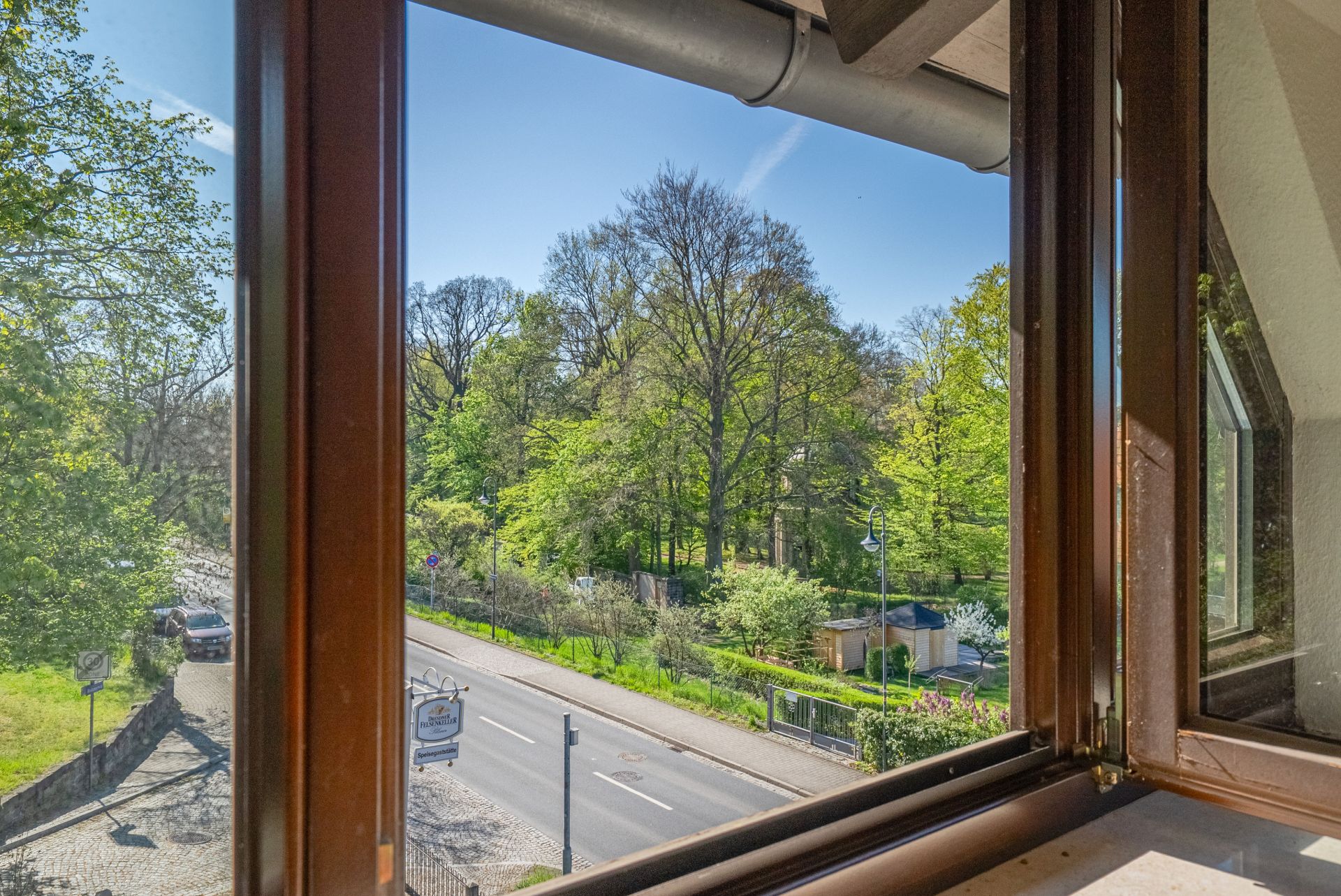 Blick aus einem geöffneten Fenster auf eine Straße und einen grünen Park mit Bäumen.