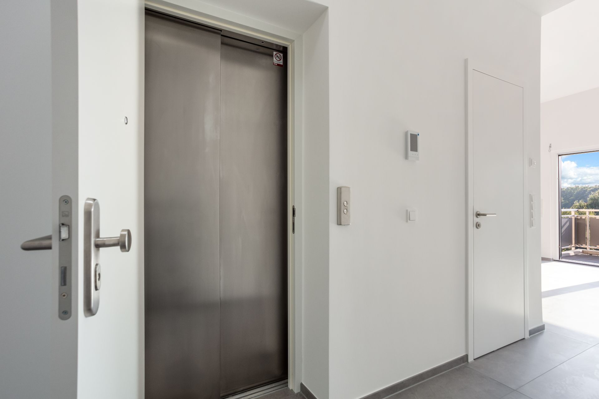 Heller Flur mit Aufzug, weißer Tür und Zugang zu einem Balkon.
