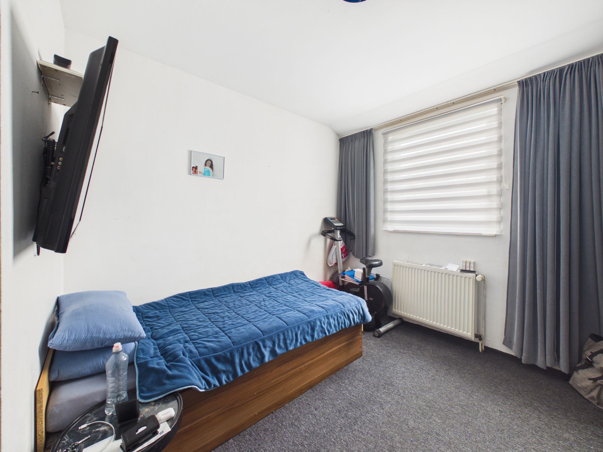 Kleines Schlafzimmer mit Einzelbett, Fernseher an der Wand und Fitnessgerät am Fenster.