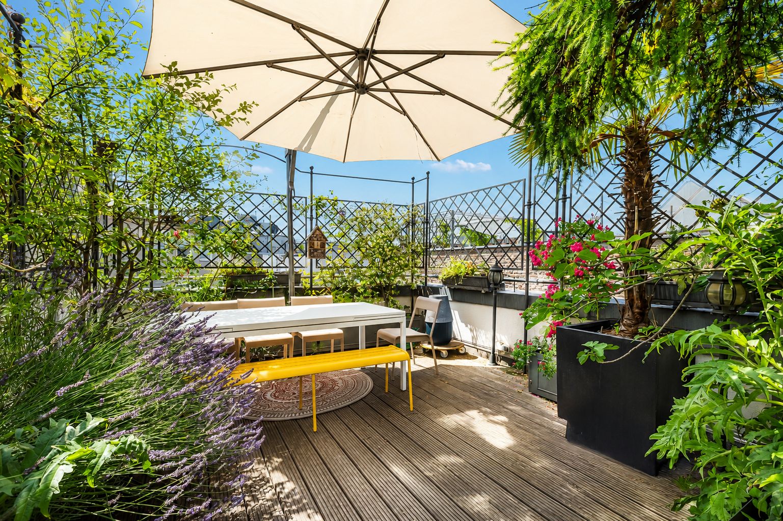 Holzterrasse mit Tisch, gelber Bank, Stühlen, Pflanzen und großem Sonnenschirm.