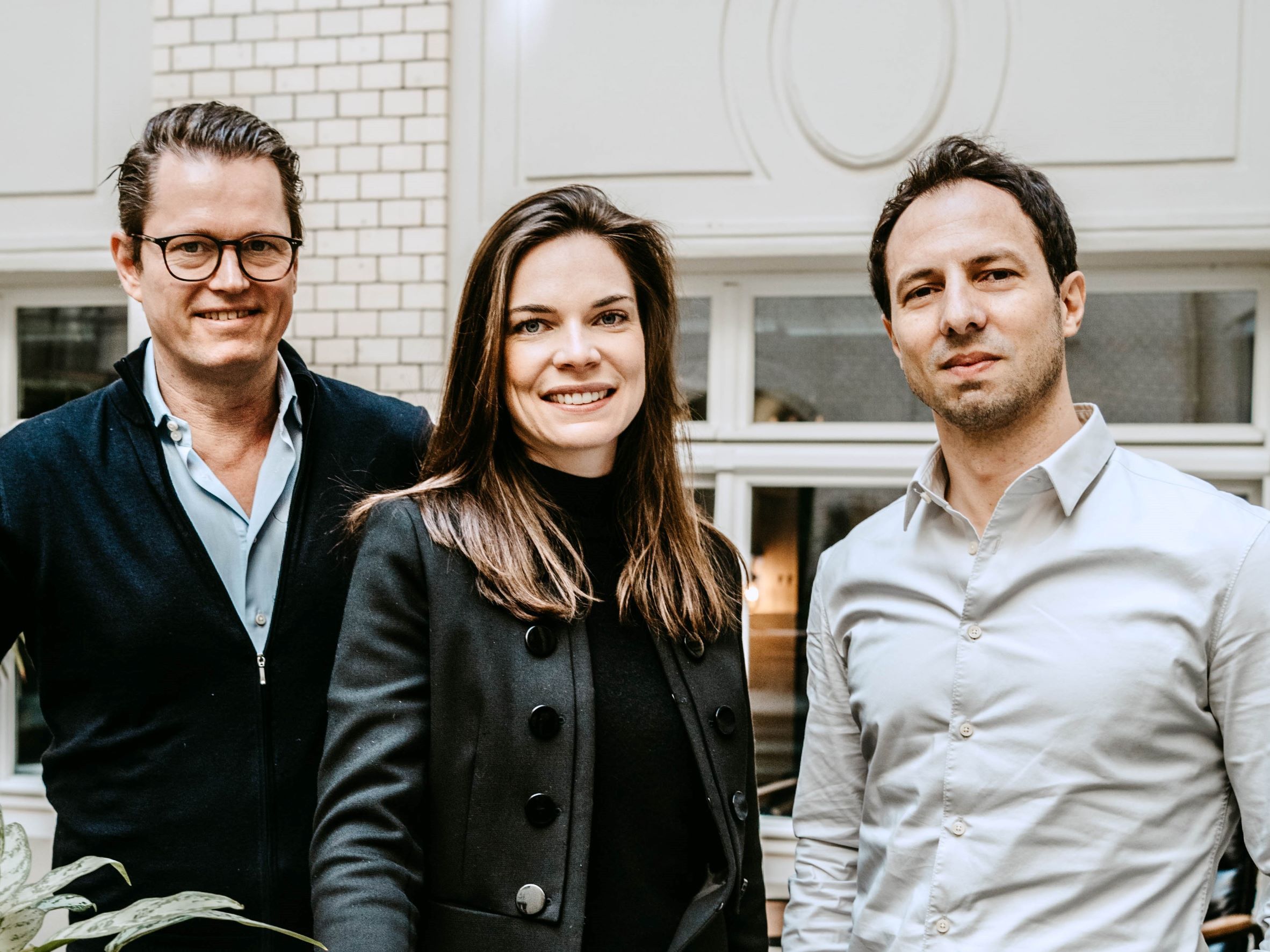 Drei Menschen stehen lächelnd in einem Innenraum vor einem Fenster mit weißem Rahmen.