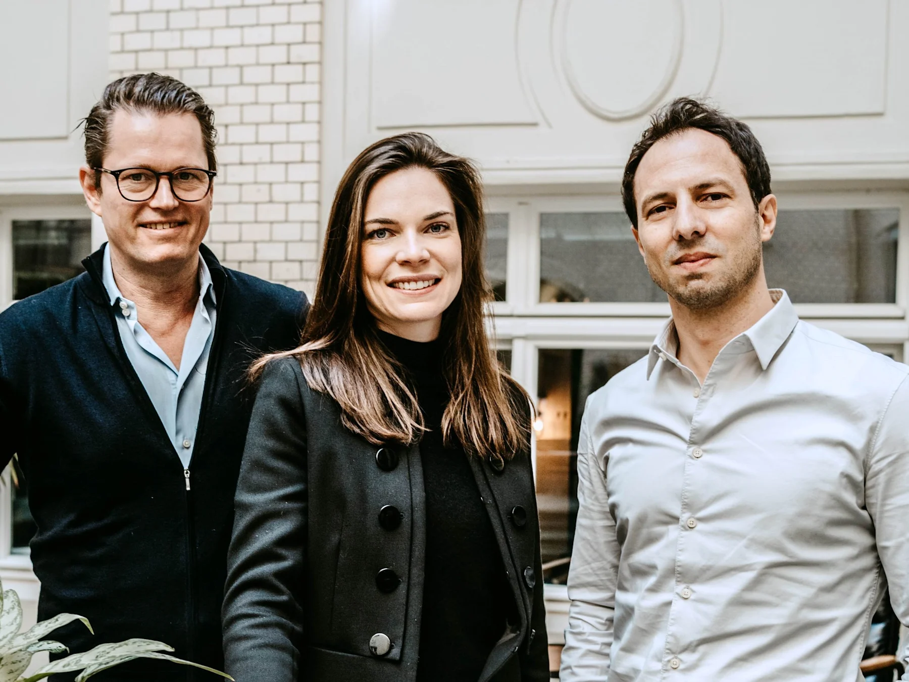 Drei Menschen stehen lächelnd in einem Innenraum vor einem Fenster mit weißem Rahmen.