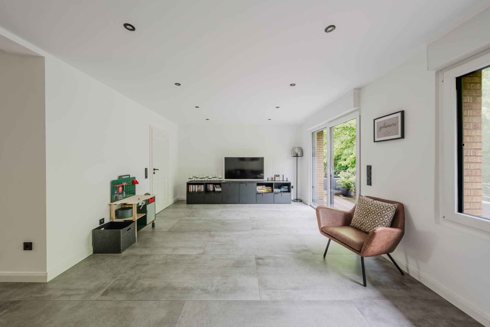 Helles Wohnzimmer mit Fernseher, Sessel, Spielzeugküche und großen Fenstern zum Garten.