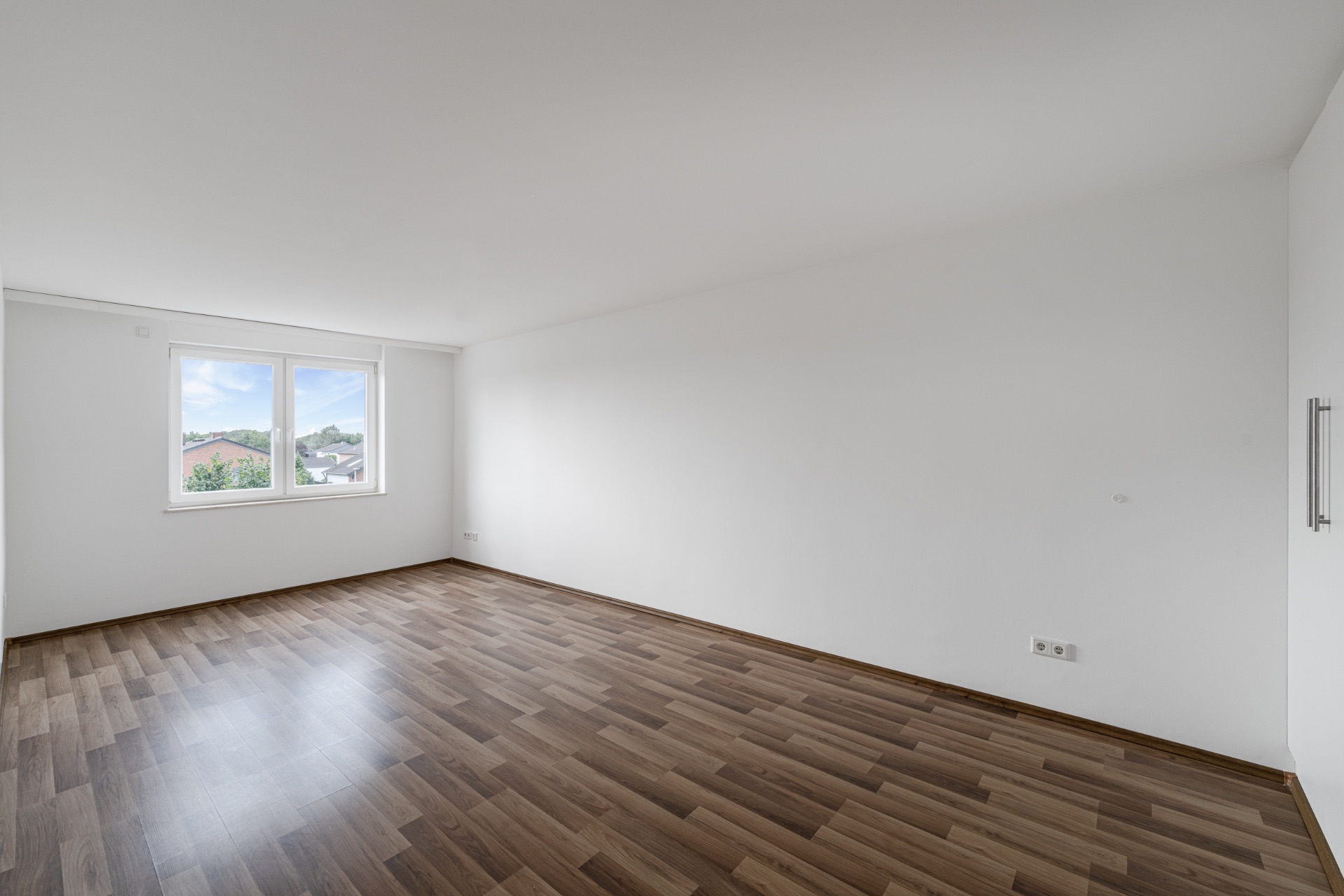 Leerer Raum mit Holzfußboden, weißen Wänden und großem Fenster mit Ausblick.