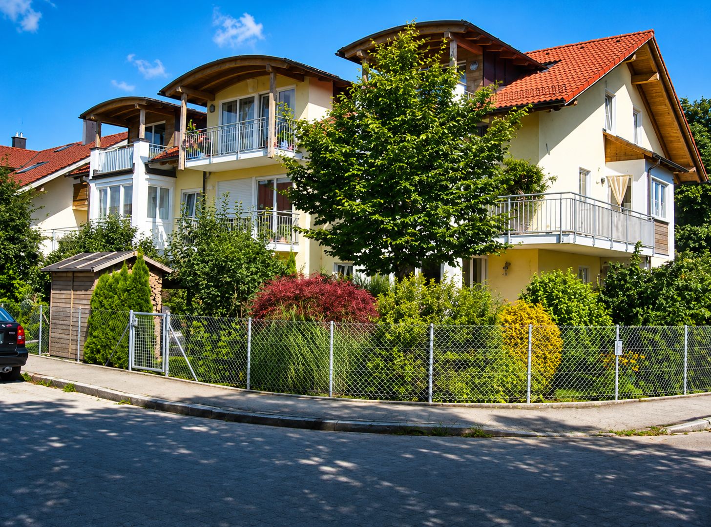 Mehrfamilienhaus mit Balkonen, Garten, Bäumen und Zaun an einer ruhigen Straße.