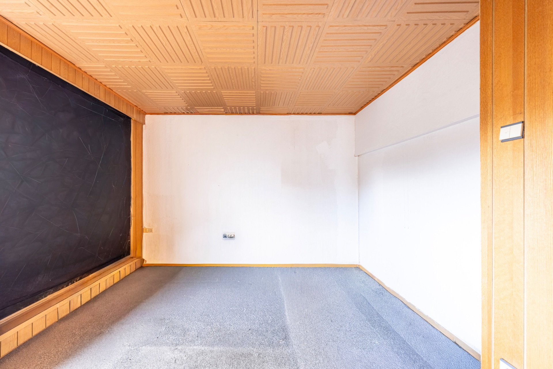 Leerer Raum mit Holzwänden, Teppichboden und großer schwarzer Tafel an der linken Wand.