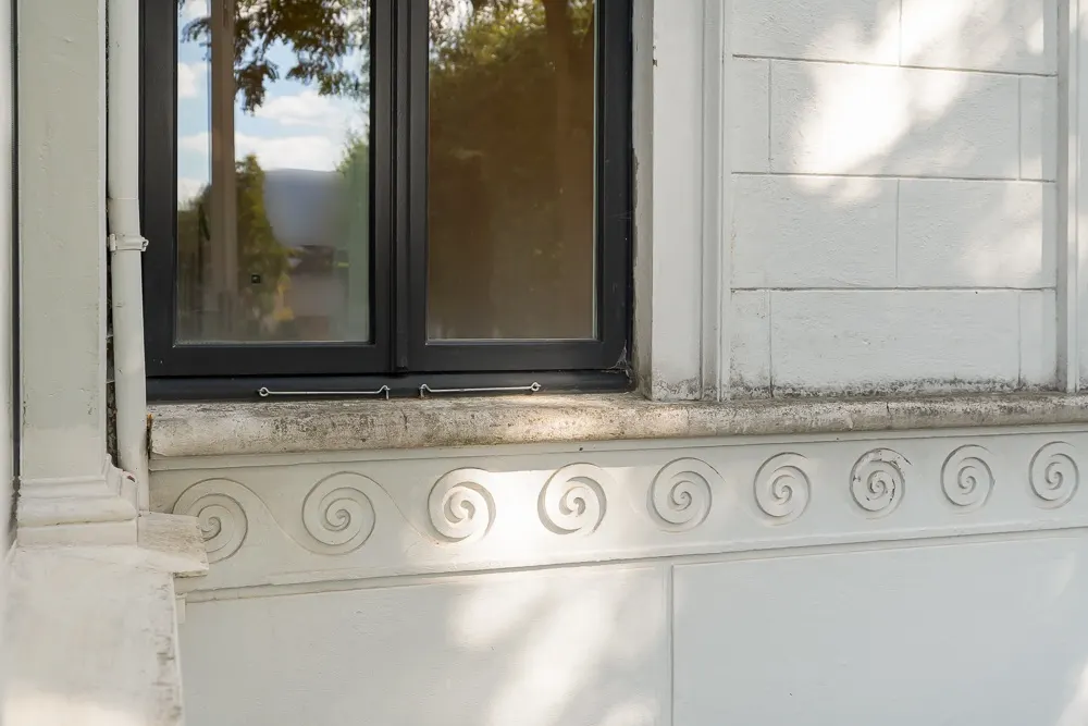Fenster mit dunklem Rahmen und verzierten weißen Stuckelementen an einer Hausfassade.