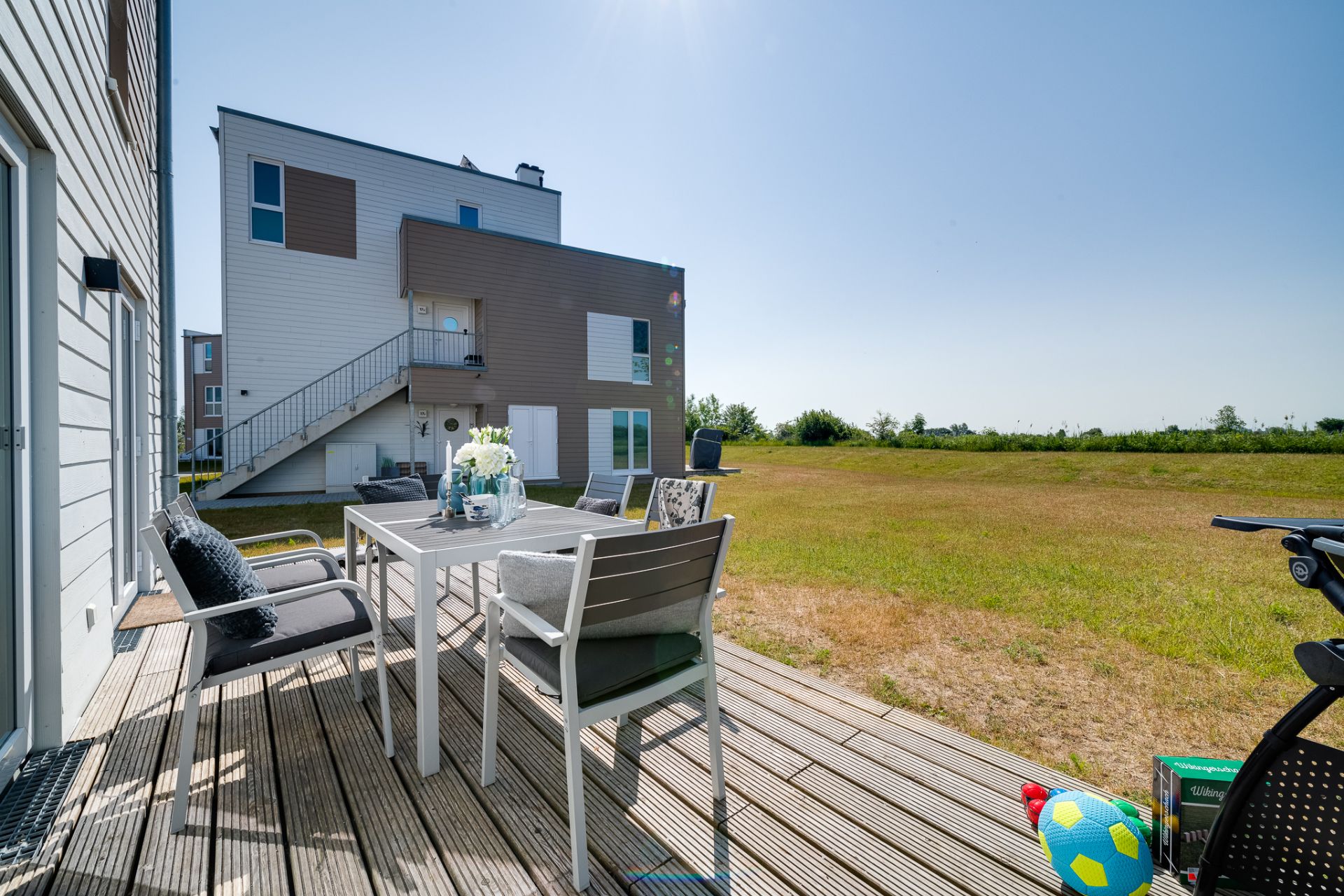 Terrasse mit Esstisch und Stühlen, Spielzeugball, moderner Wohnblock im Hintergrund, Wiese im Sonnenschein.