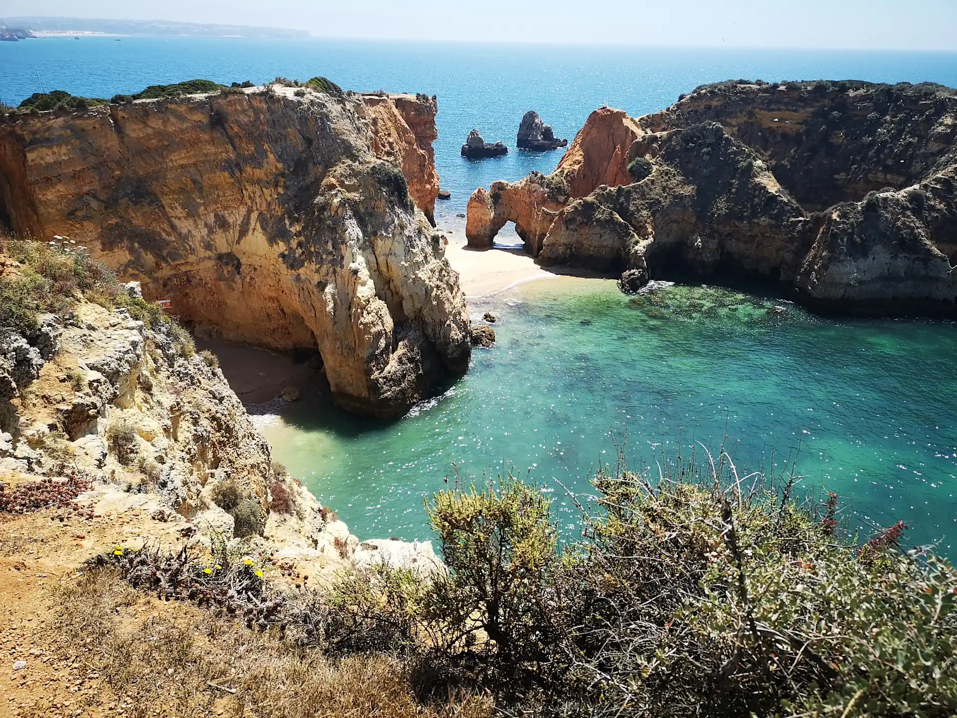 Klippen mit Blick auf türkisfarbenes Meer und Sandstrand, im Vordergrund trockene Pflanzen.