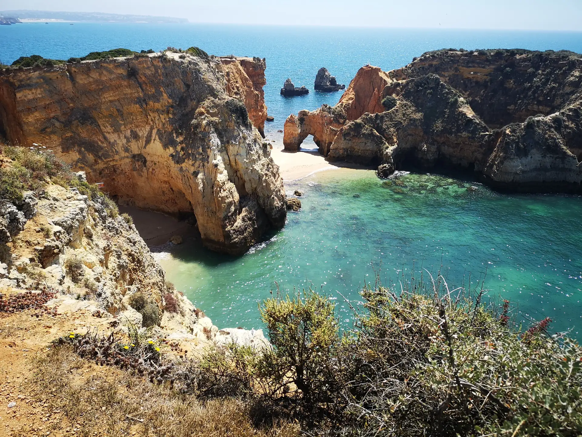 Klippen mit Blick auf türkisfarbenes Meer und Sandstrand, im Vordergrund trockene Pflanzen.