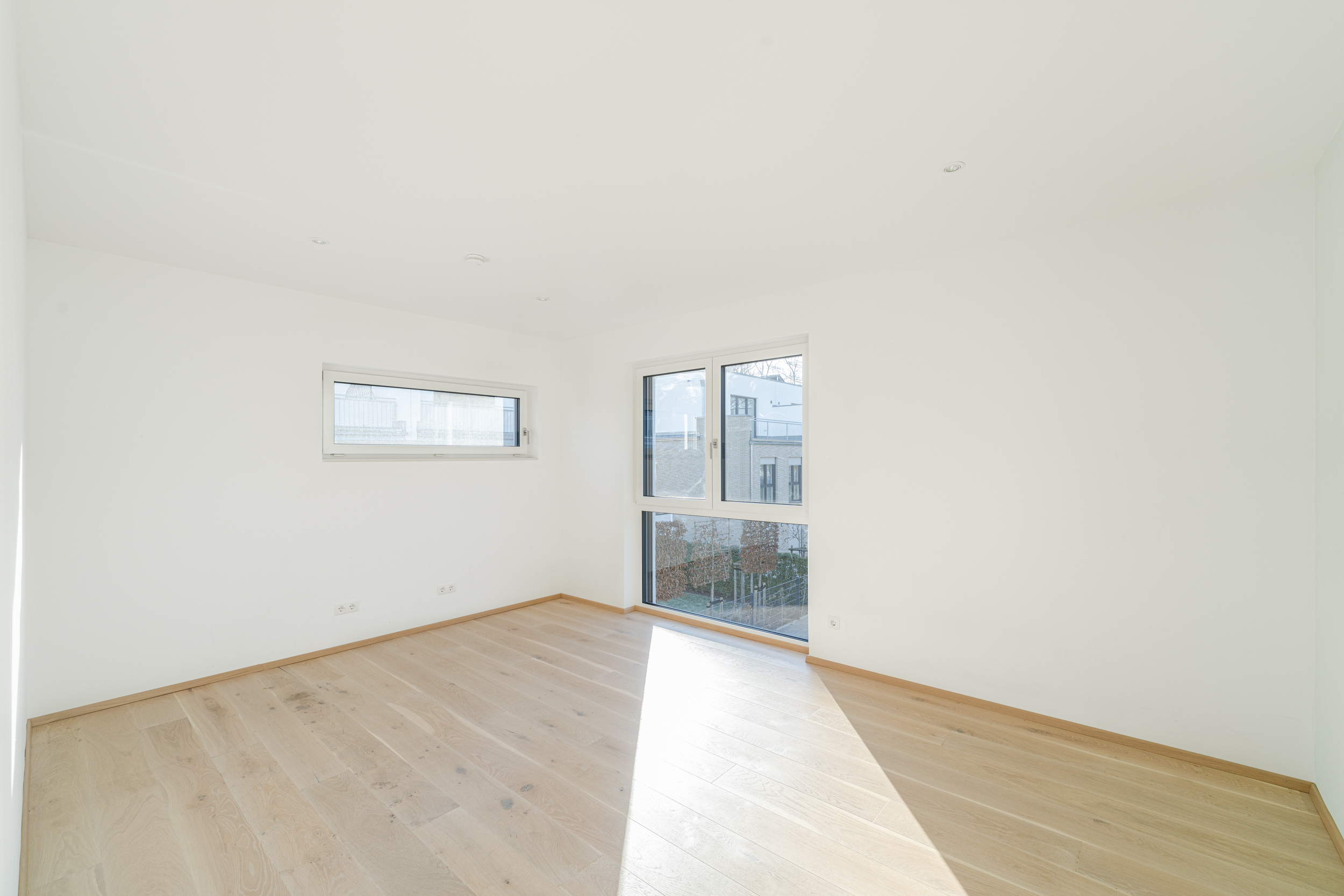 Leerer, heller Raum mit Holzfußboden und zwei großen Fenstern.
