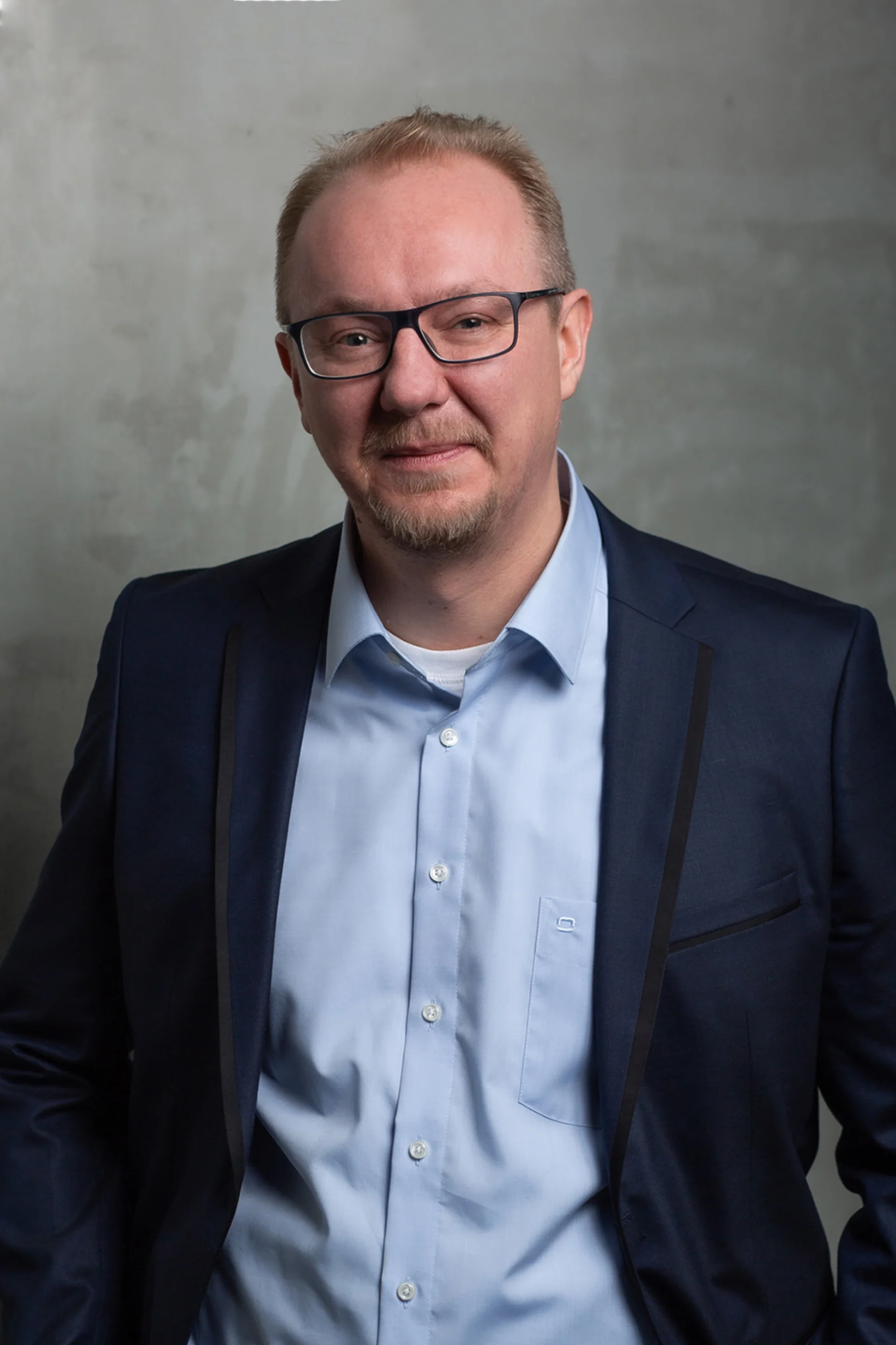 Ein Mann mit Brille und Bart trägt ein blaues Hemd und ein dunkles Sakko vor grauem Hintergrund.