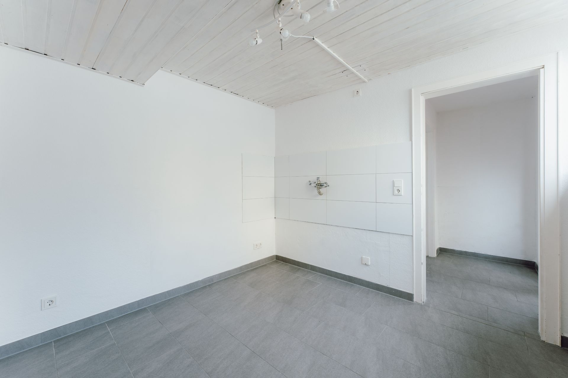 Leerer Raum mit grauem Fliesenboden, weißen Wänden und Decke, Wasseranschluss an der Wand.