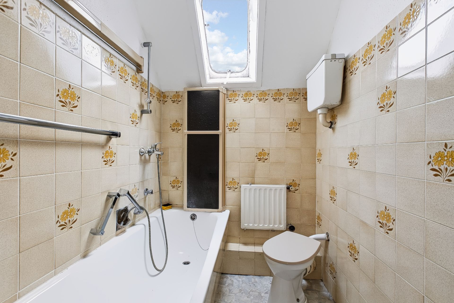 Kleines Badezimmer mit Badewanne, Toilette, Heizkörper und Dachfenster, beige Fliesen mit Blumenmuster.