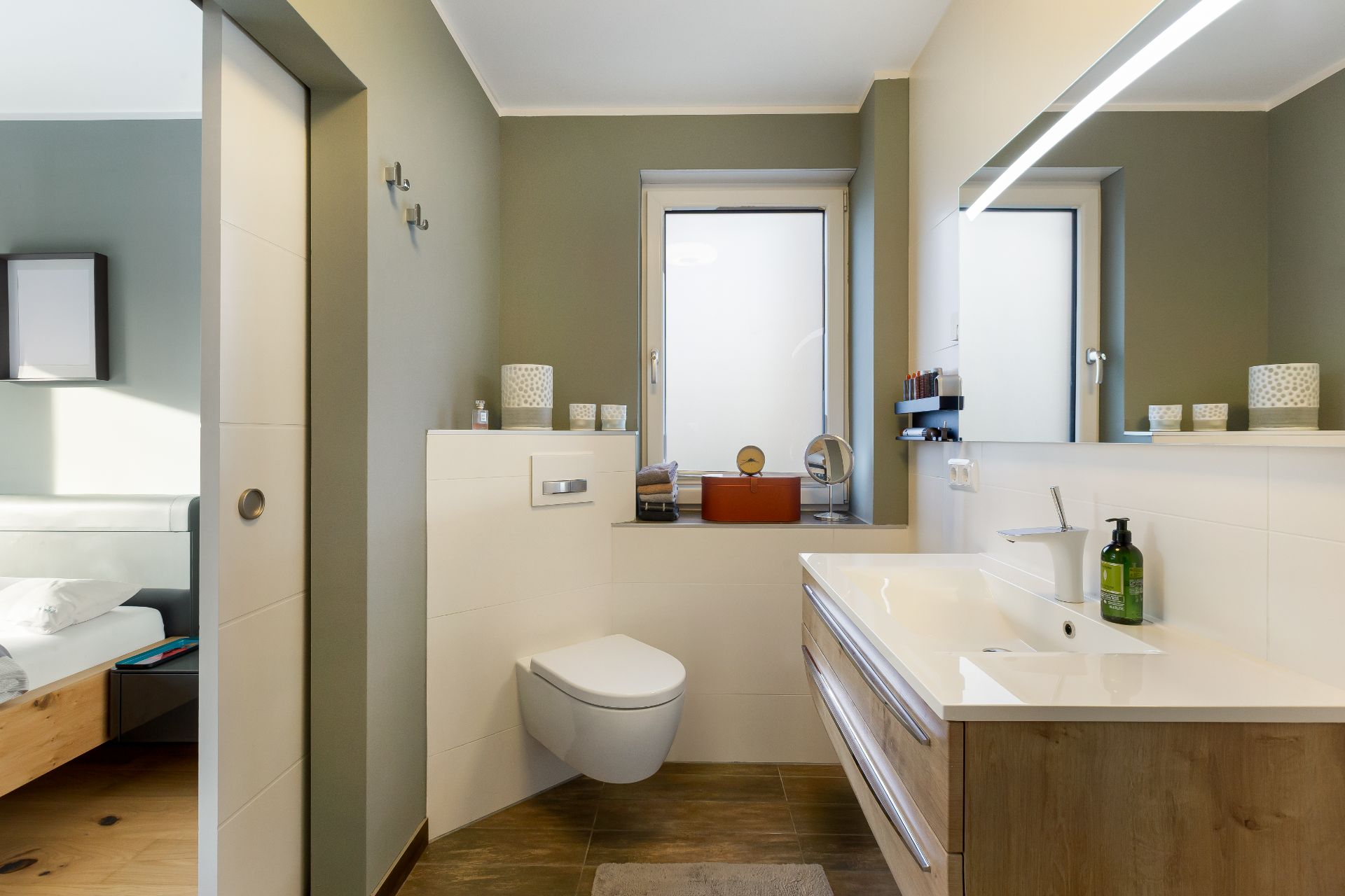 Modernes Badezimmer mit Waschbecken, Spiegel, Toilette und Fenster, angrenzendes Schlafzimmer sichtbar.