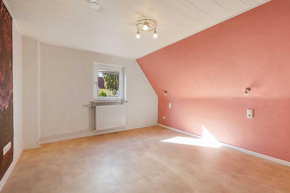 Helles Zimmer mit schräger, rosa Wand, Fenster und Deckenleuchte.